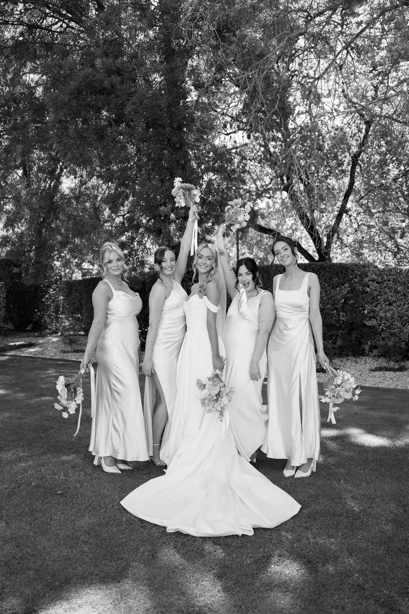 Black and white photo of five women in bridesmaid dresses celebrating outdoors, holding bouquets, with trees in the background.