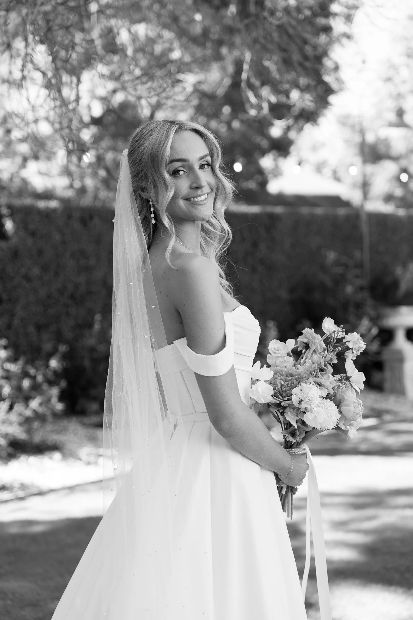 A bride in a white wedding gown with off-the-shoulder sleeves, holding a bouquet of flowers, outdoor setting with trees and a bench in the background, black and white photo.