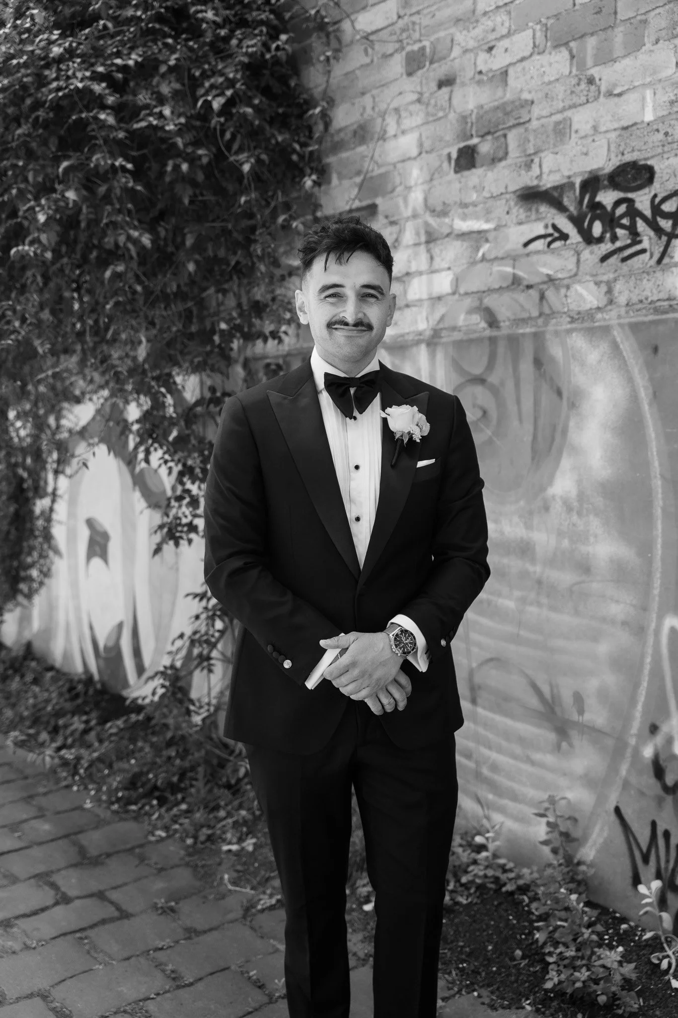 A man in a tuxedo and bow tie standing outdoors against a brick wall with graffiti, smiling, holding a rolled-up paper, wearing a wristwatch, with greenery in the background.