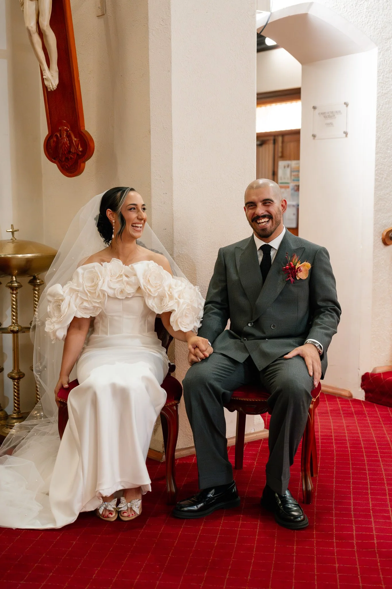 A bride and groom sitting on chairs, holding hands and smiling during a wedding ceremony. The bride is wearing a white dress with large floral embellishments on the shoulders and a veil, and the groom is wearing a gray suit with a black tie and bouto