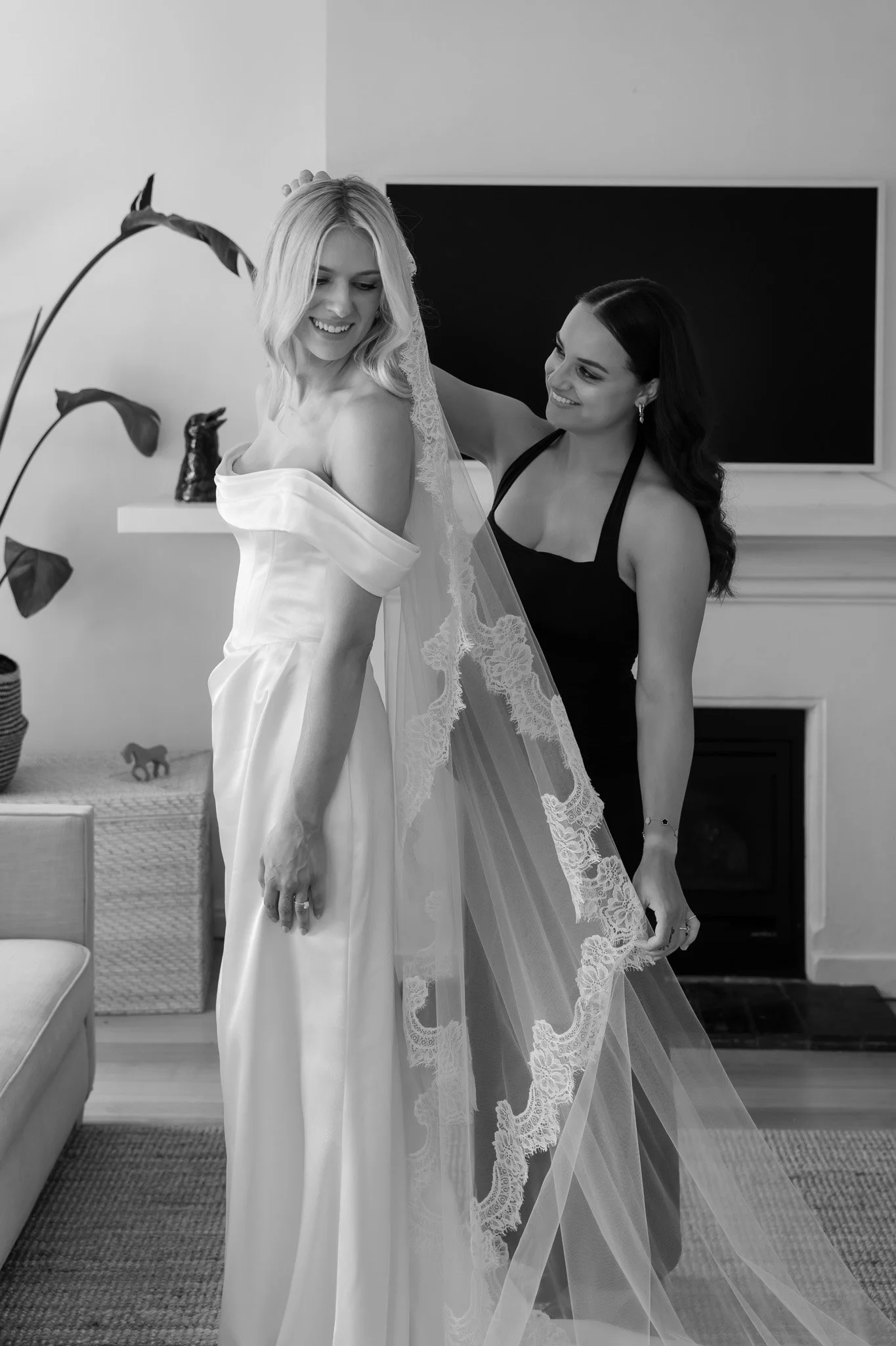 A bride in a wedding dress with a veil being assisted by a woman in a black dress, in a domestic setting with a TV and fireplace.