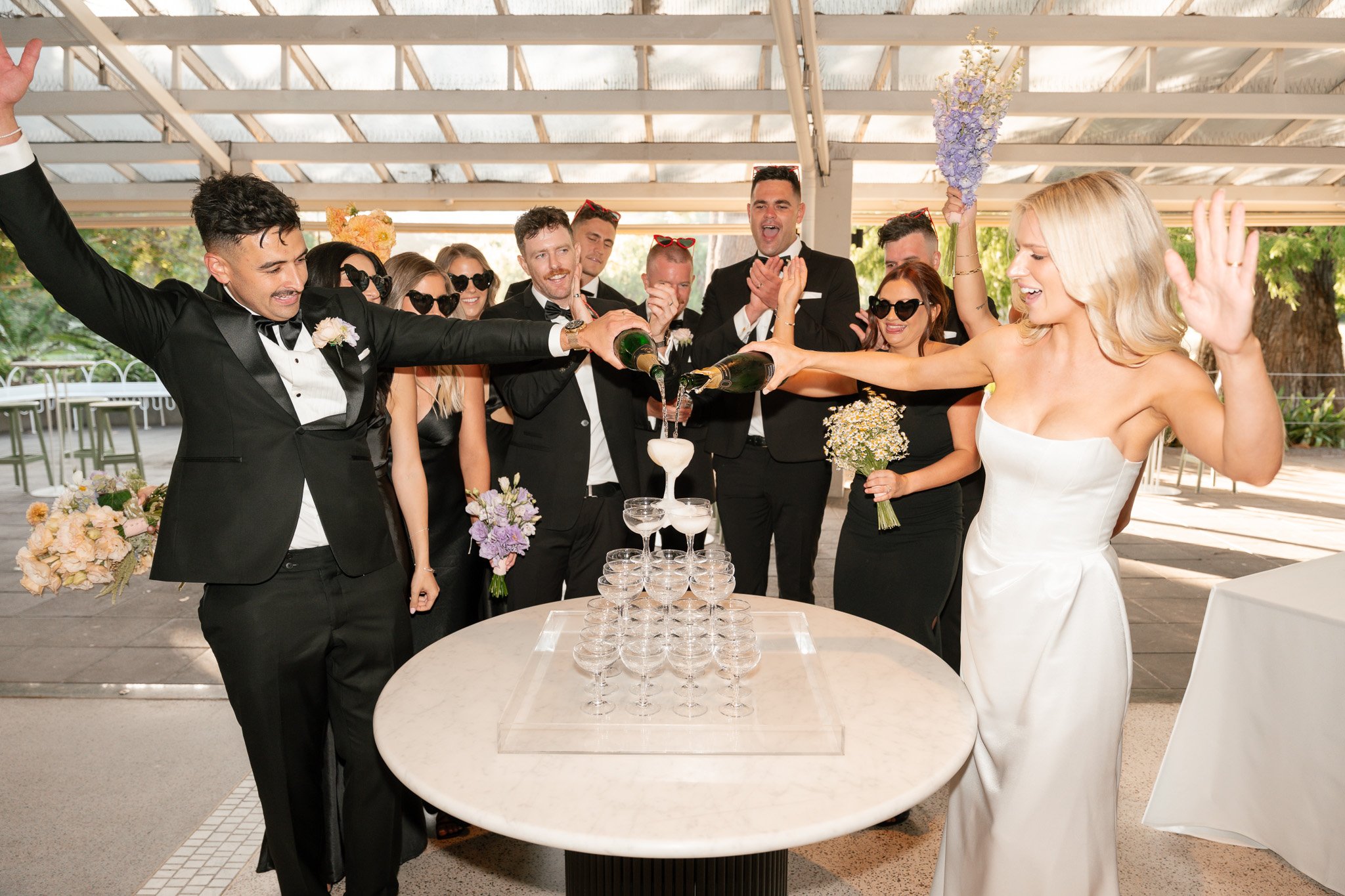 Wedding celebration with a bride and groom pouring champagne into a pyramid of glasses, surrounded by friends in formal attire.