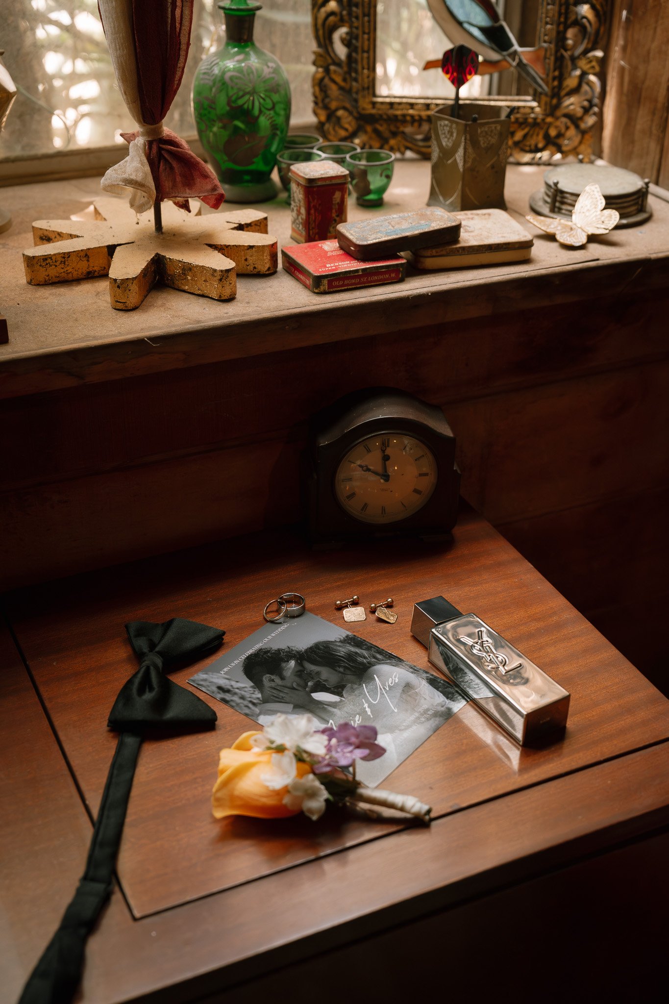 A wooden desk with a black clock, wedding rings, a black bow tie, flowers, and a wedding invitation, with a mostly wooden and decorative background.