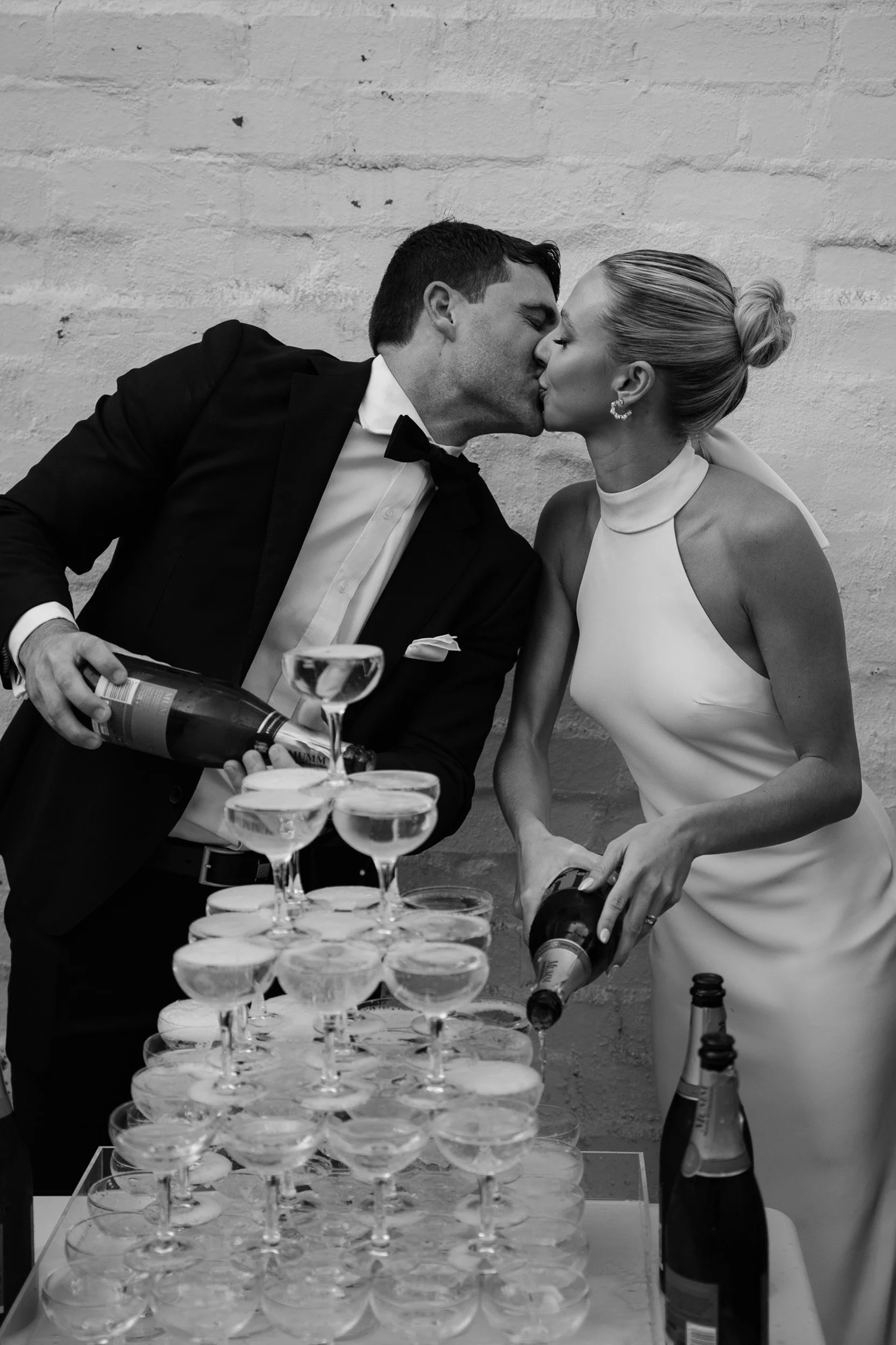 A black and white photo of a couple in wedding attire kissing while pouring champagne into a tower of glasses.