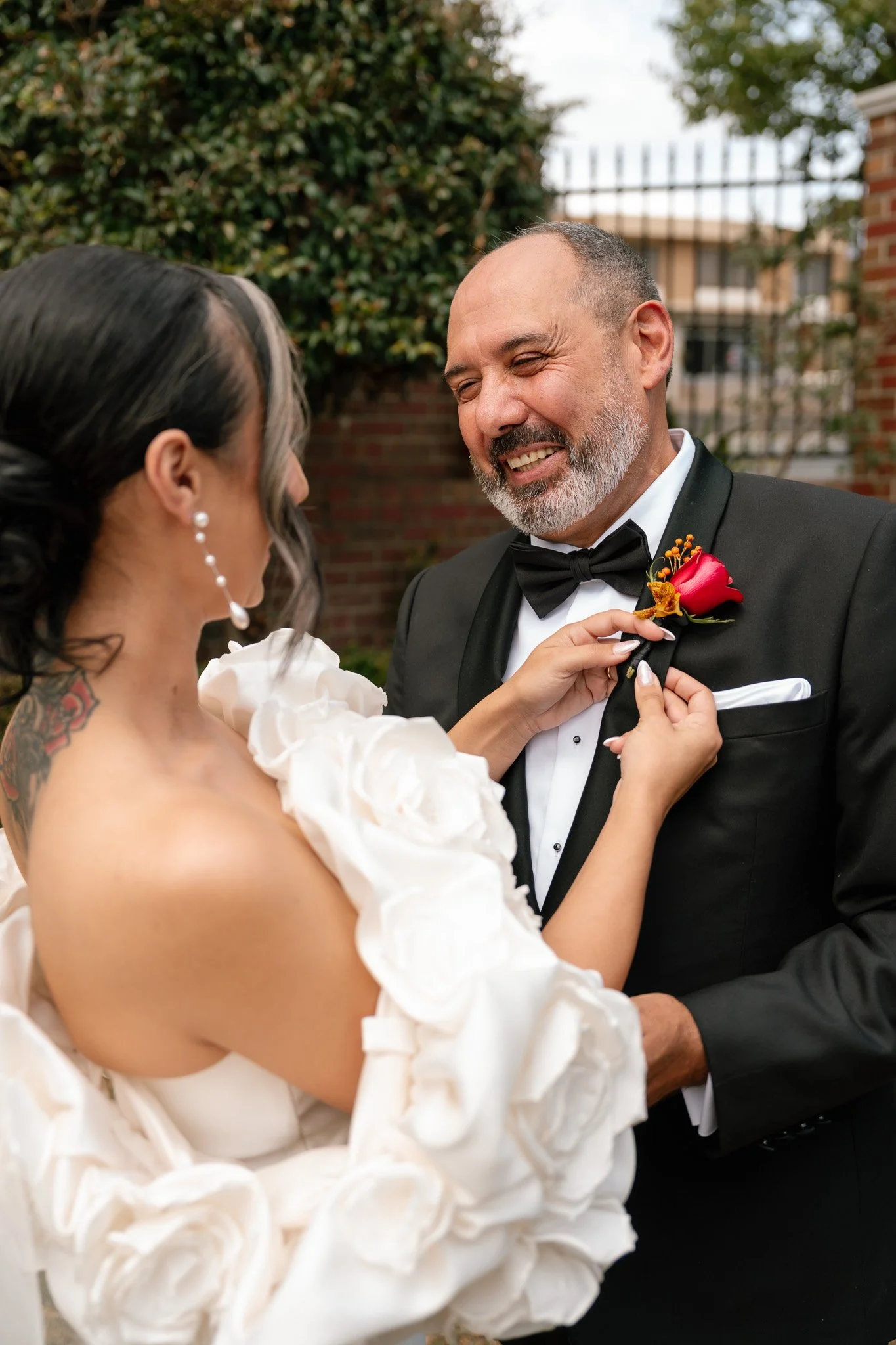 A bride pinning a red boutonniere on a groom's black tuxedo jacket outdoors, with trees and a brick wall in the background.