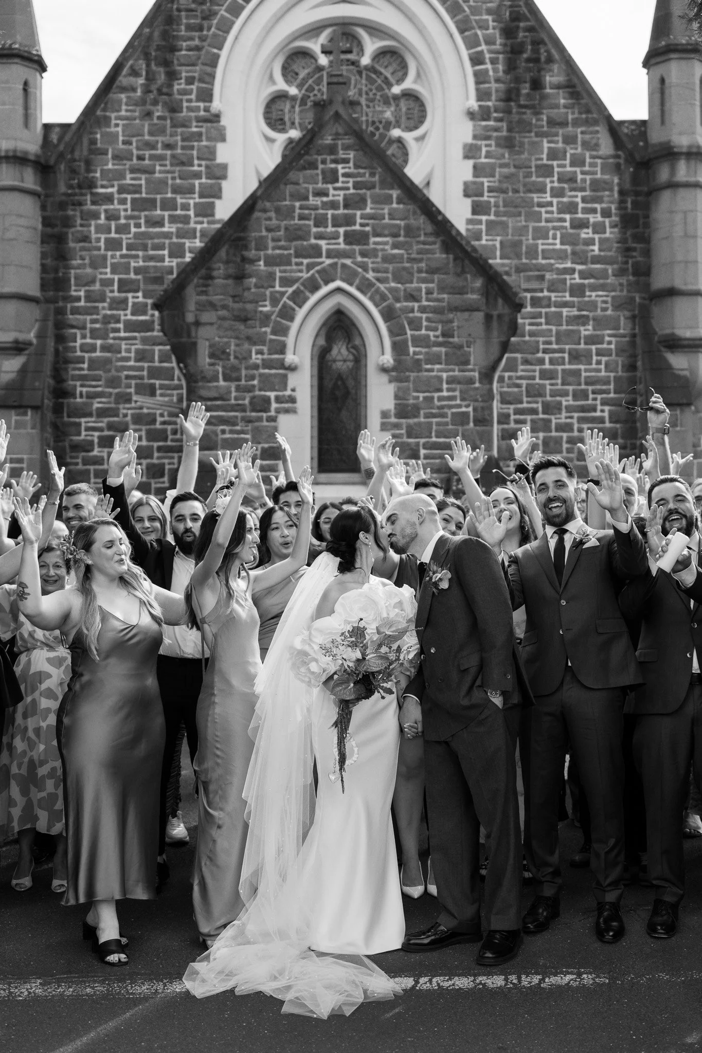 A black and white photo of a wedding celebration outside a church, with a bride and groom kissing in front of friends and family.