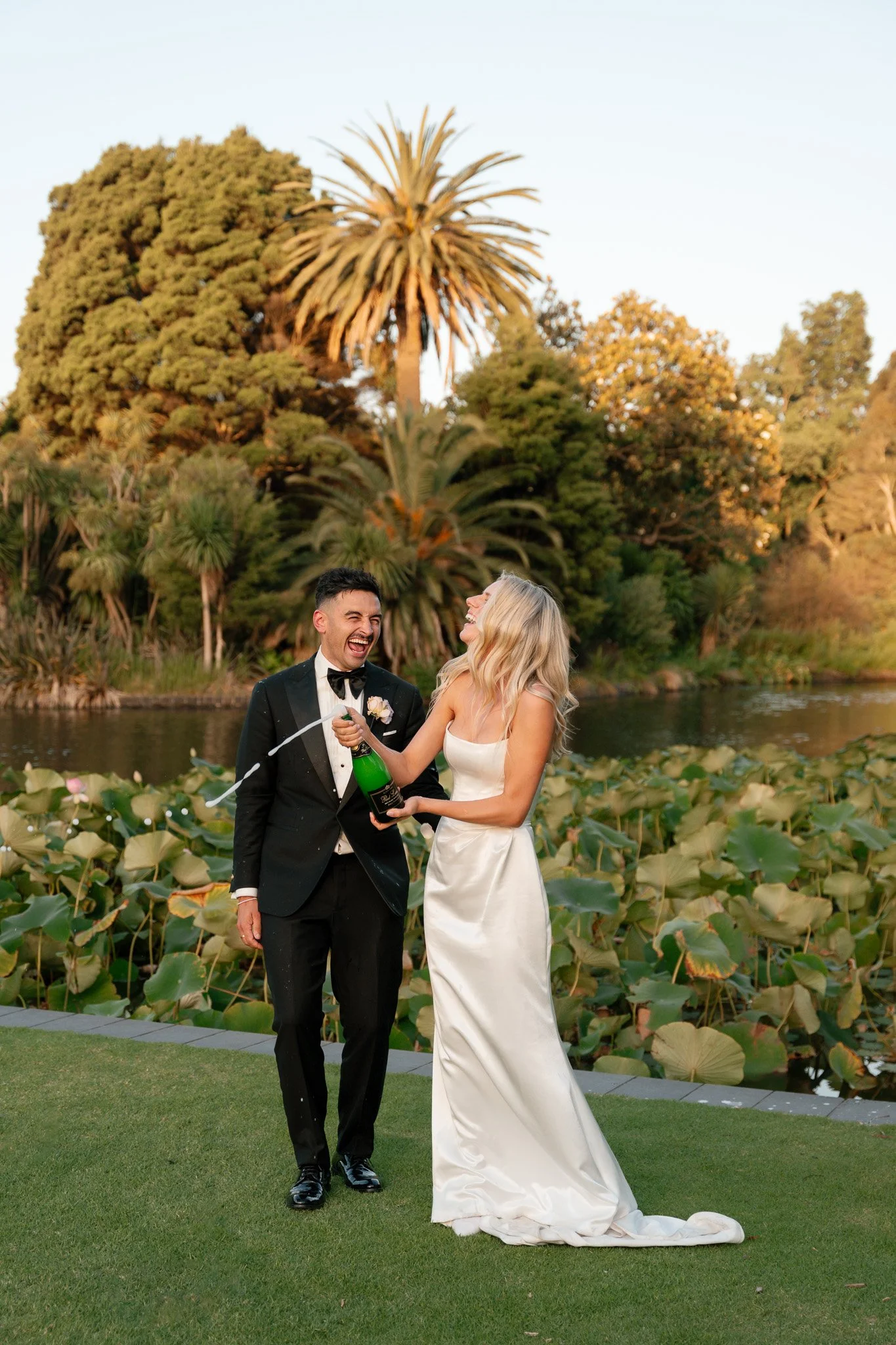 A newlywed couple celebrating outdoors near a pond with water lilies, palm trees, and lush greenery during sunset, with one person opening a champagne bottle.