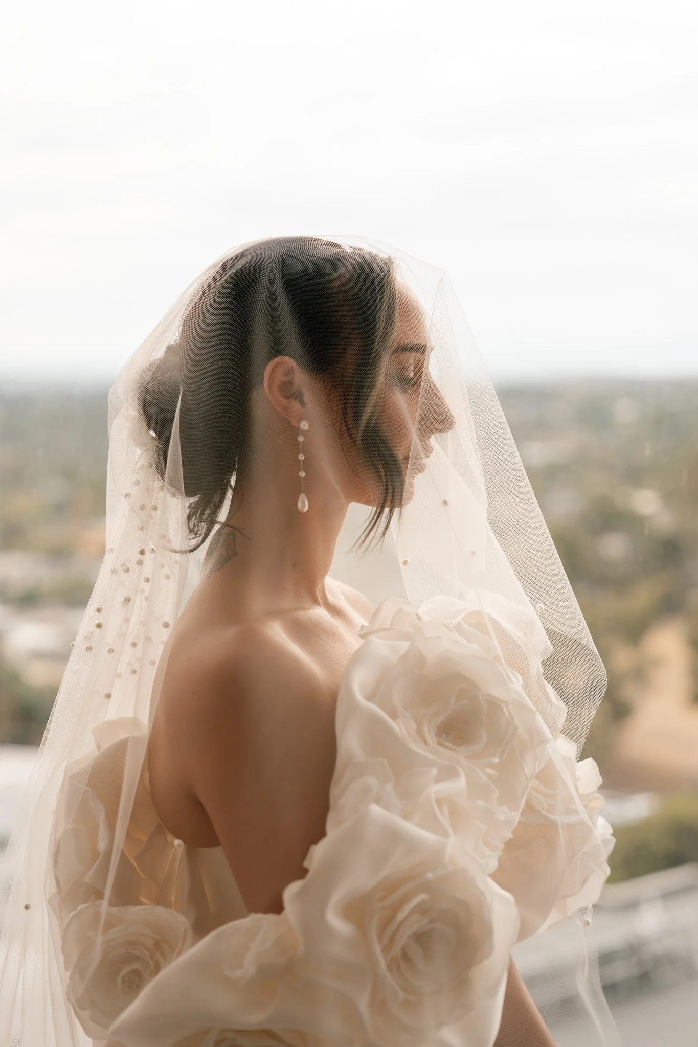 Bride with dark hair, wearing a veil and pearl earrings, gazing down with a serious expression, surrounded by white fabric resembling roses.