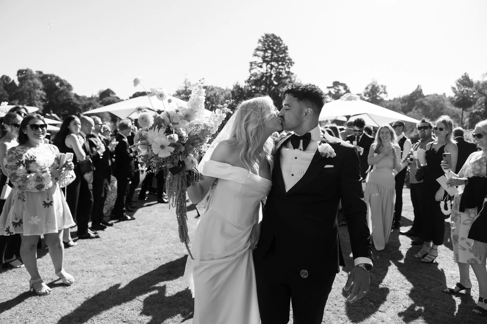 A black and white photo of a newlywed couple sharing a kiss outdoors, surrounded by wedding guests under umbrellas.