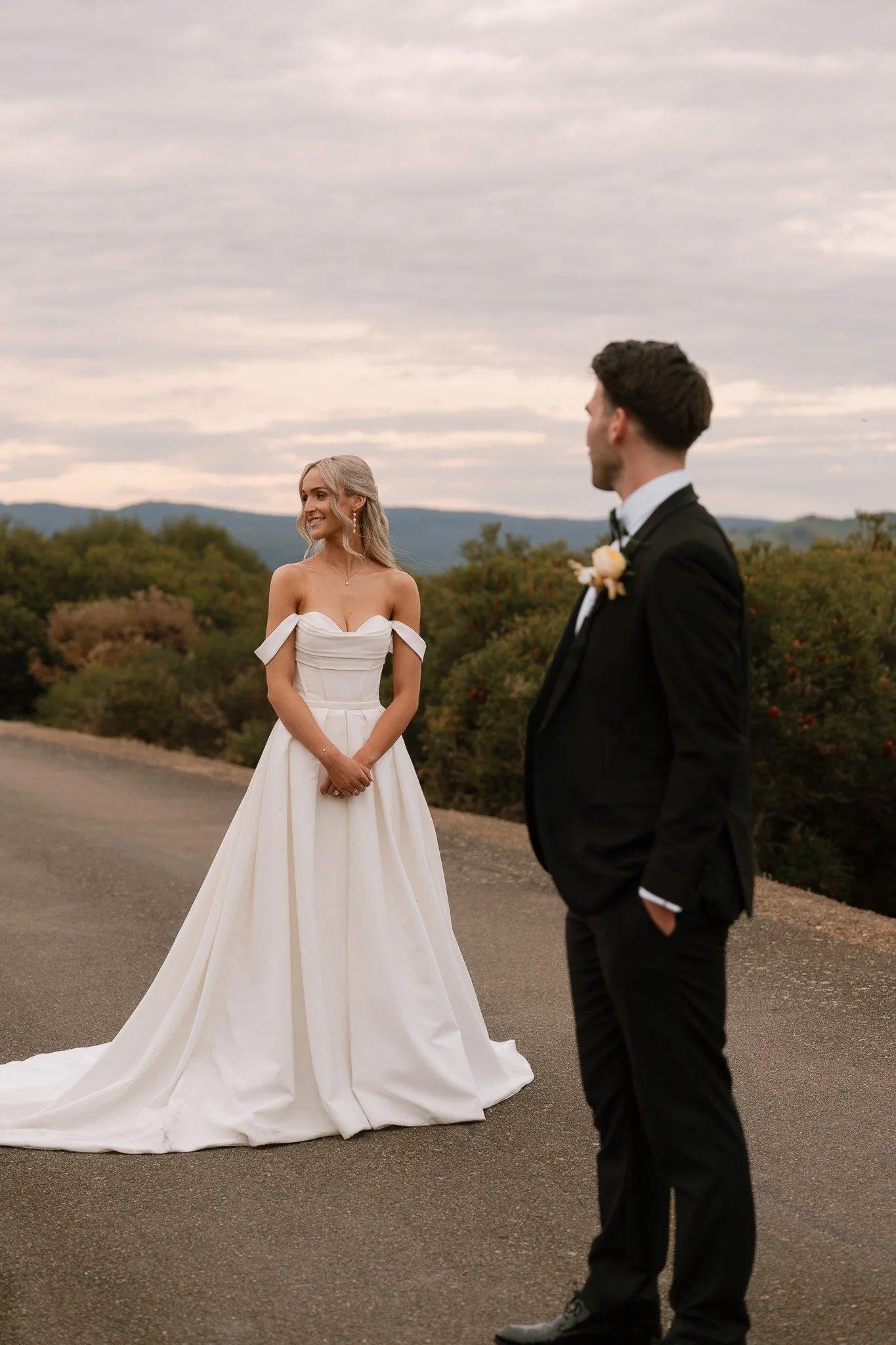 Bride in a white wedding dress and groom in a black suit standing outdoors with a scenic background and cloudy sky.