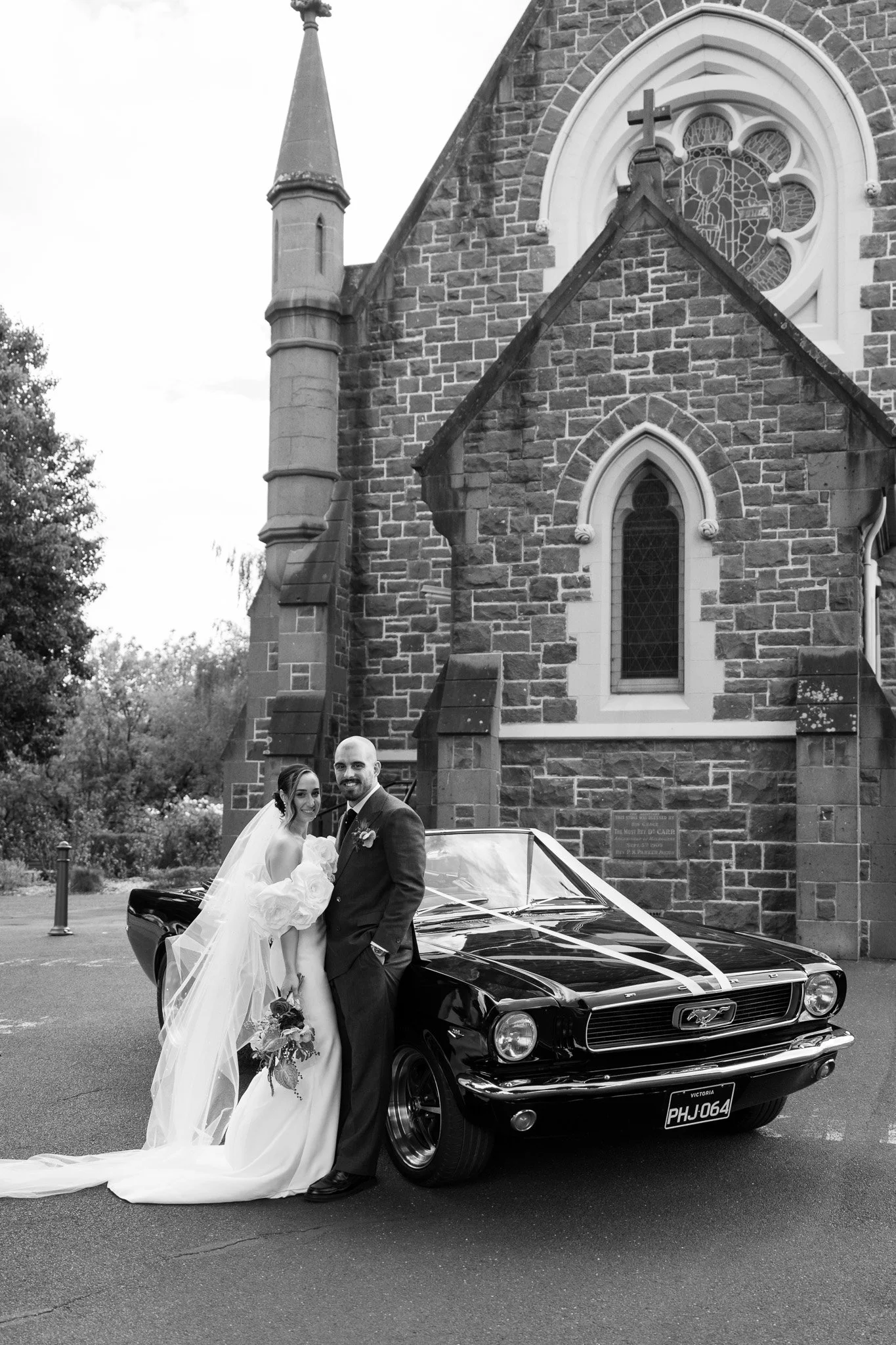 A bride and groom standing next to a vintage black Ford Mustang with a white ribbon on the hood, in front of a stone church, celebrating their wedding day.