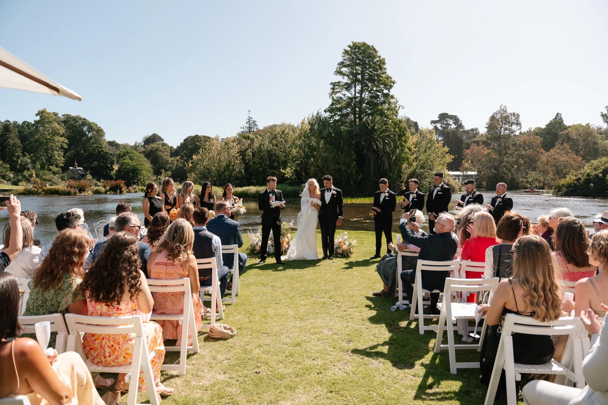 Outdoor wedding ceremony by a river with the bride and groom in the center, officiant reading, bridal party standing nearby, guests seated on either side, green trees and blue sky in the background.
