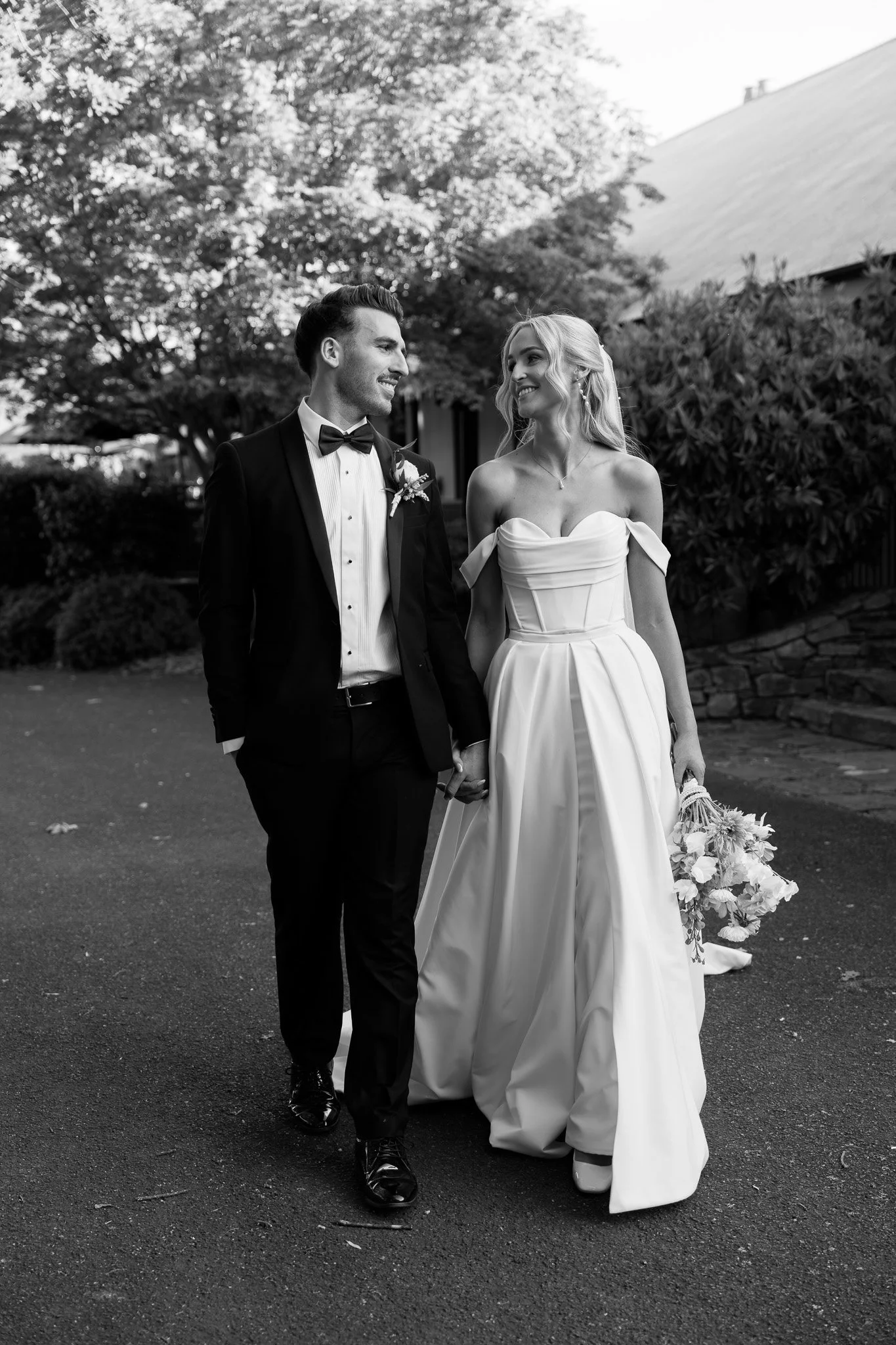 A bride and groom walking outside on their wedding day, holding hands and smiling at each other. The bride is wearing a strapless wedding gown and holding a bouquet, while the groom is dressed in a tuxedo with a bow tie.