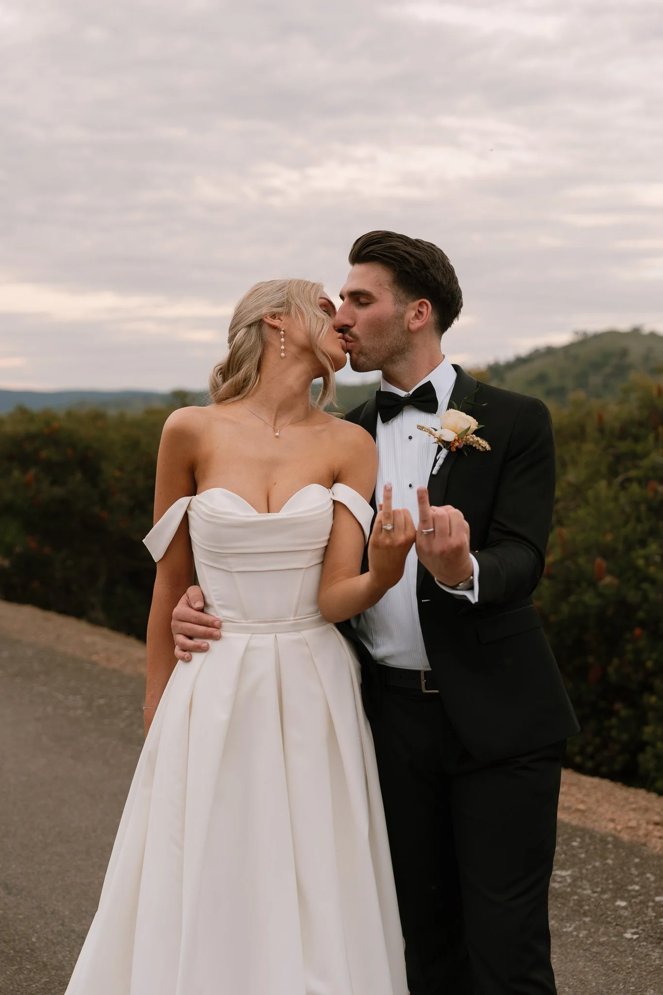 A bride and groom kiss and show their middle fingers in a scenic outdoor setting.