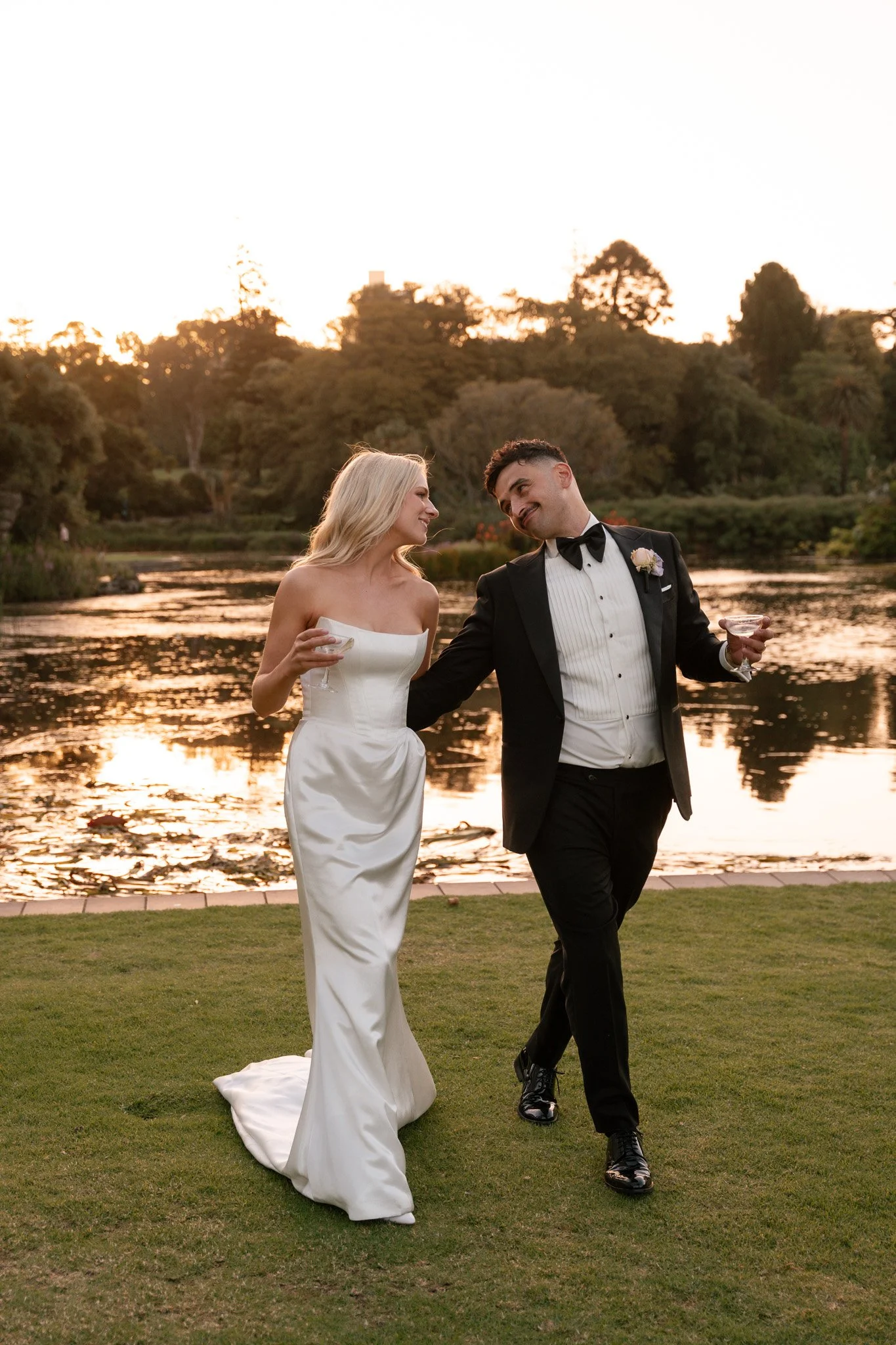 A newlywed couple walking by a lake at sunset, smiling and holding drinks, with the bride in a strapless white gown and the groom in a tuxedo.