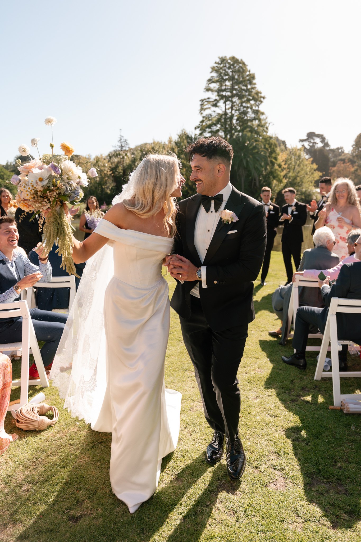 A bride and groom smiling and dancing at an outdoor wedding ceremony on a sunny day, surrounded by guests seated on chairs and standing in a lush green park with trees in the background.