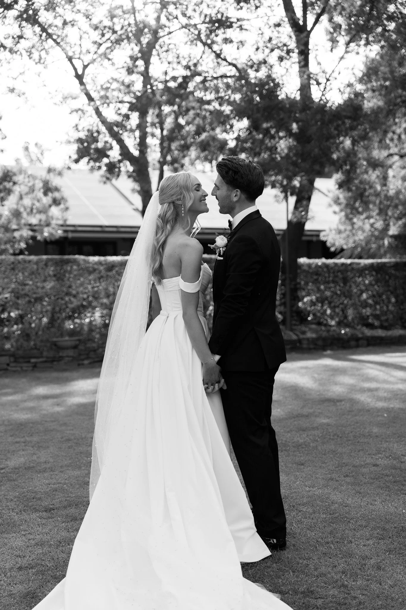 Black-and-white photo of a bride and groom holding hands, looking at each other and smiling outdoors on a wedding day.