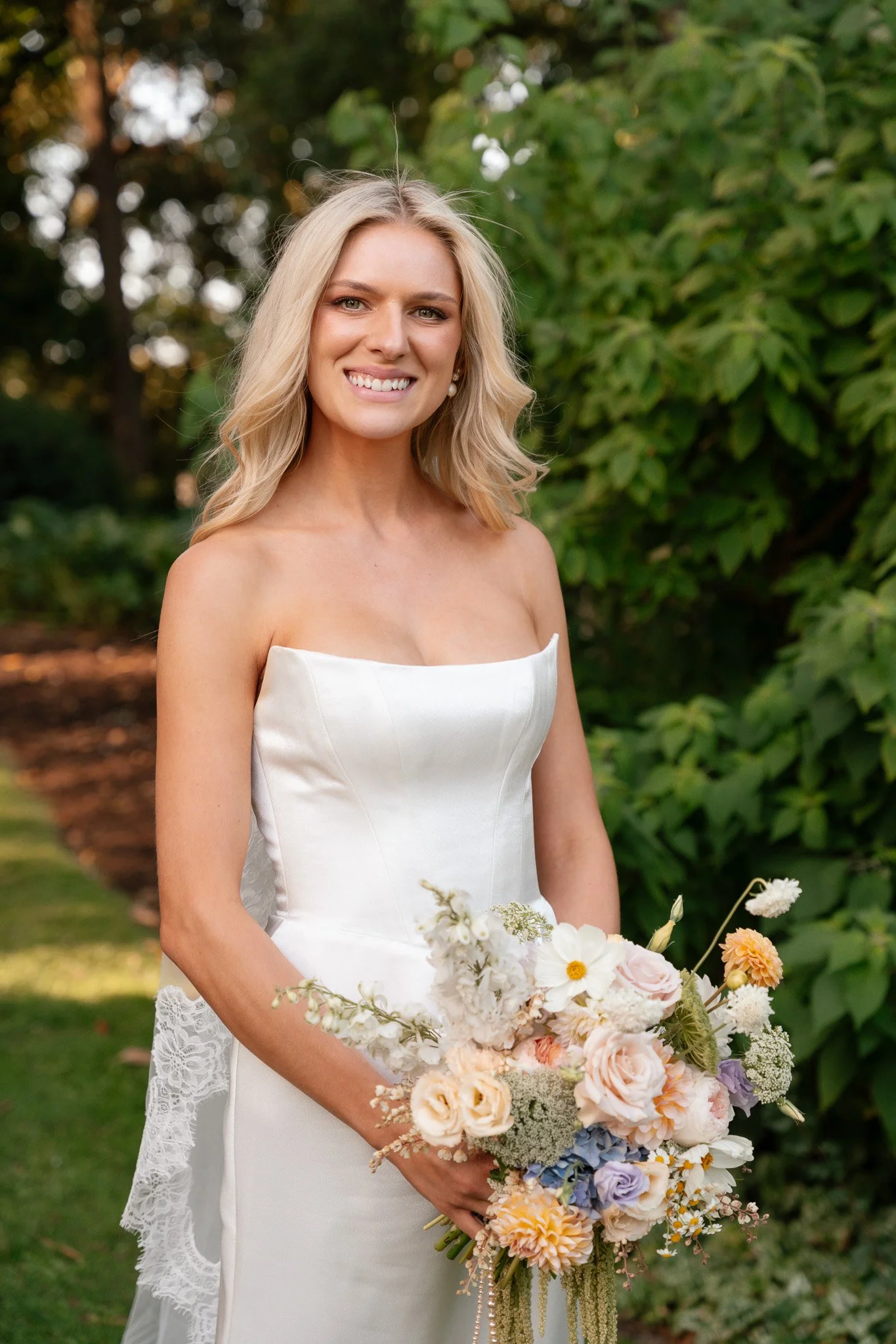 A smiling blonde woman in a white strapless wedding dress holds a colorful bouquet of flowers outdoors, with green foliage in the background.