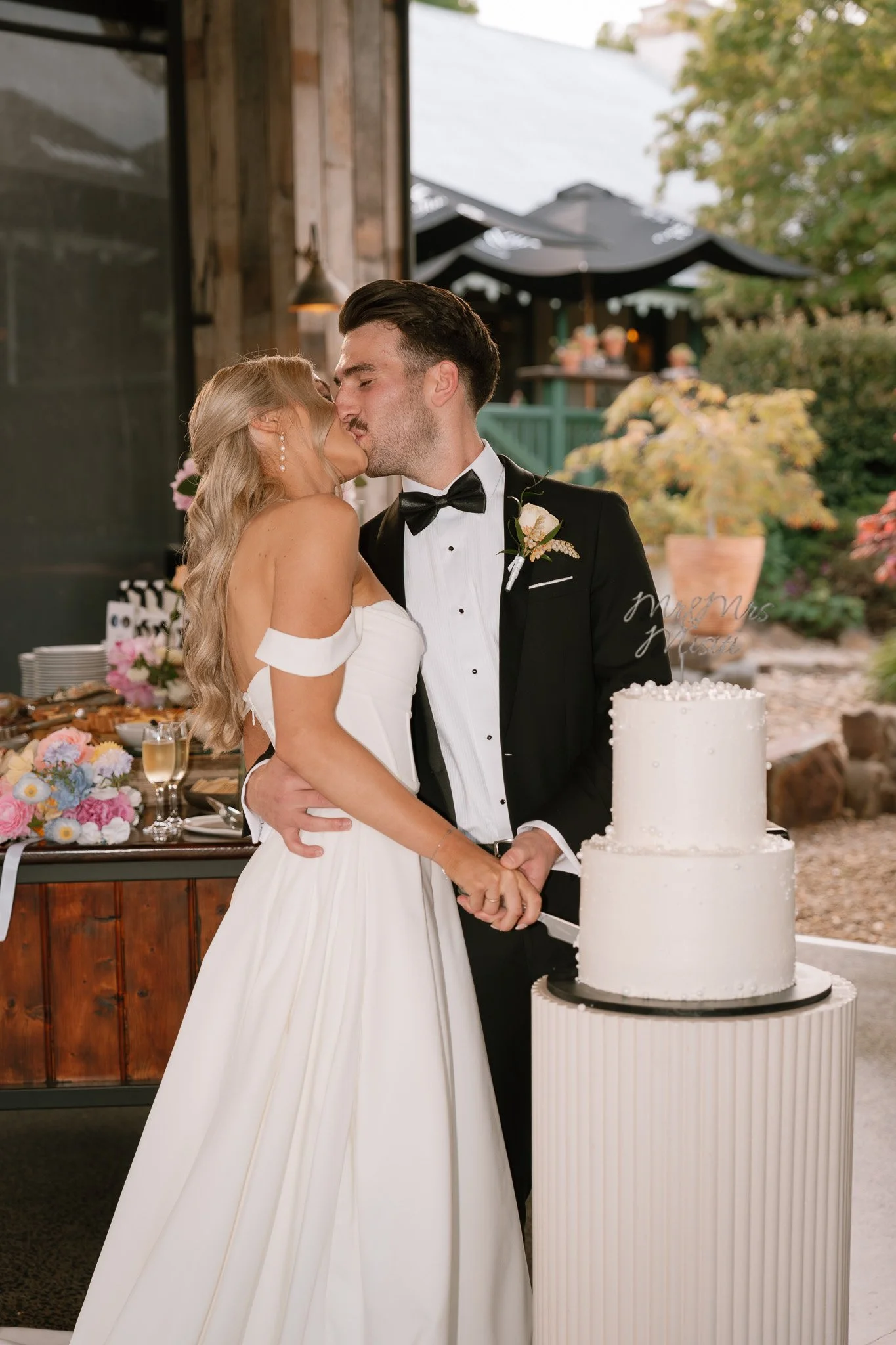 A bride and groom share a kiss while cutting their wedding cake outdoors.