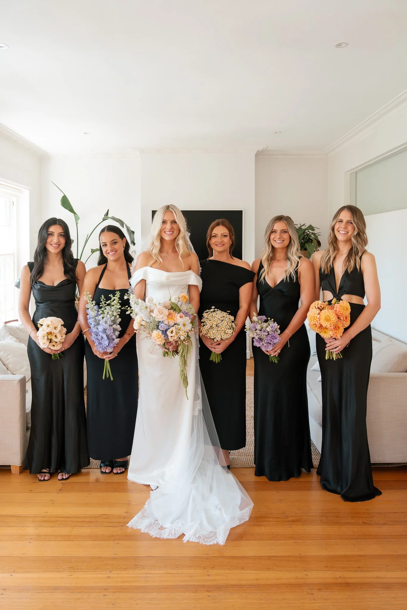 Group of six women in a living room, with a bride in a white wedding dress and five bridesmaids in black dresses, all holding bouquets of flowers.