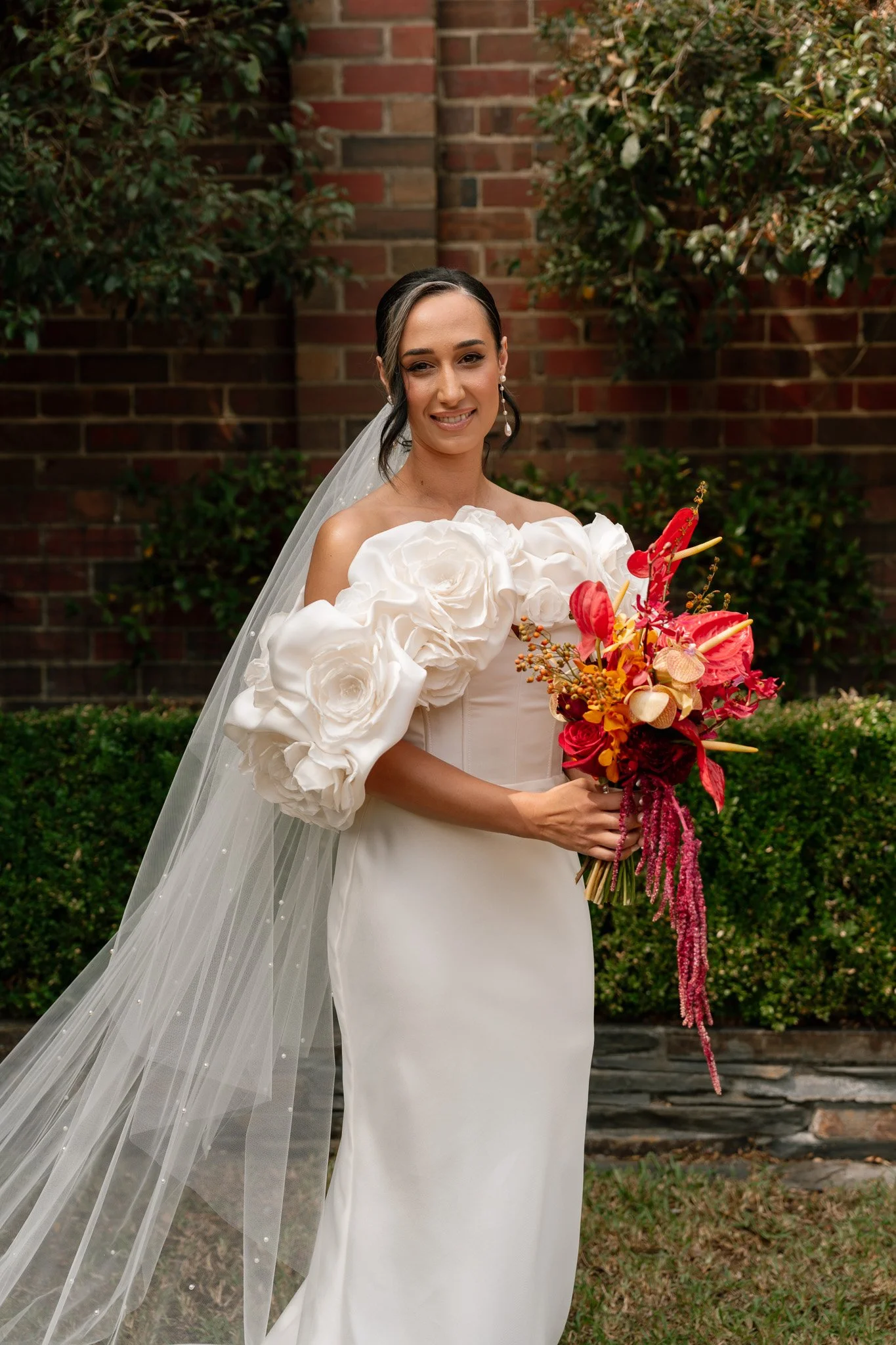 Bride in a white wedding dress holding a colorful bouquet standing outside with greenery and a brick wall in the background.