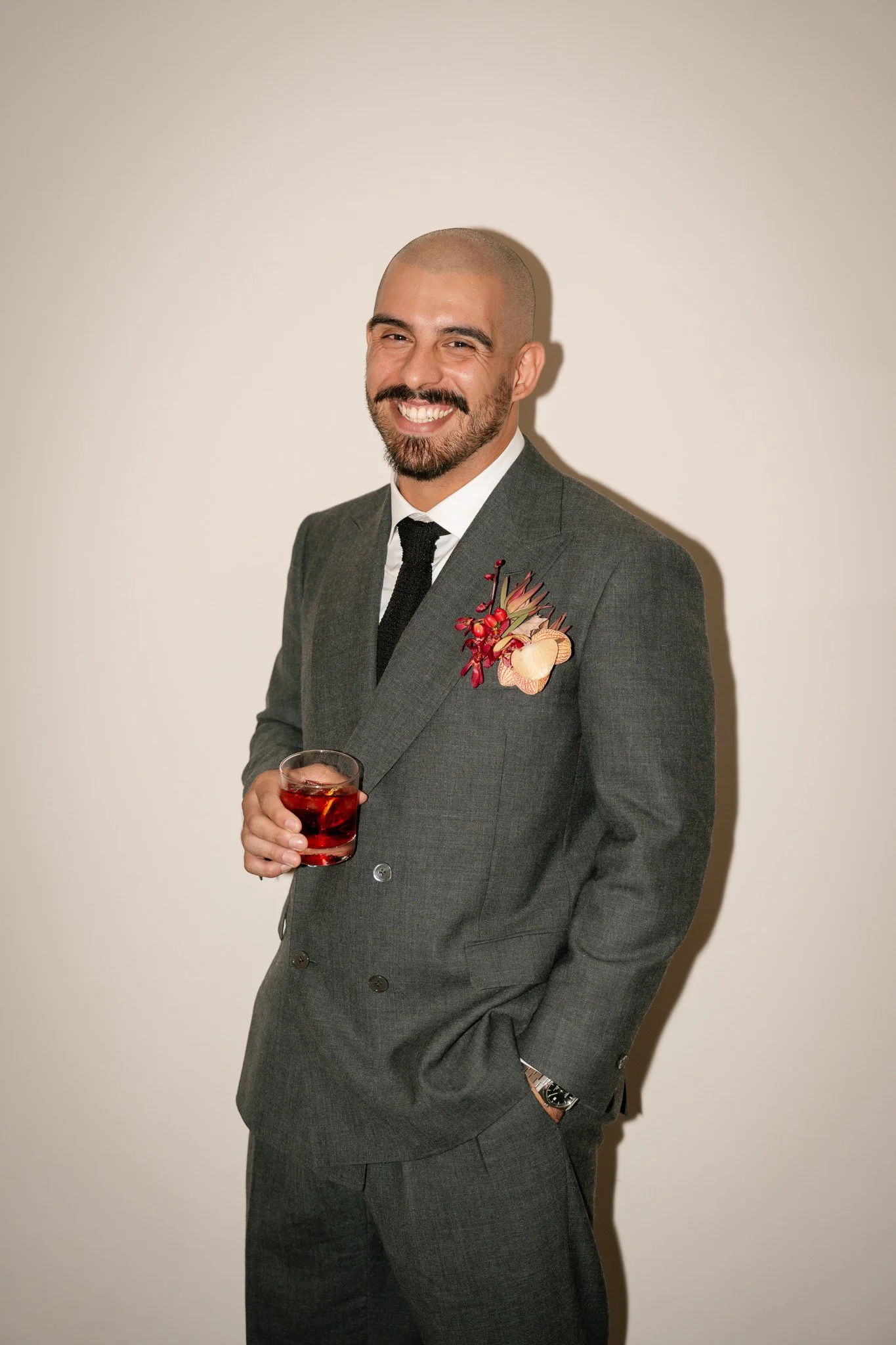 A man in a gray suit, black tie, and white shirt smiling while holding a glass of red drink, standing against a plain white wall.