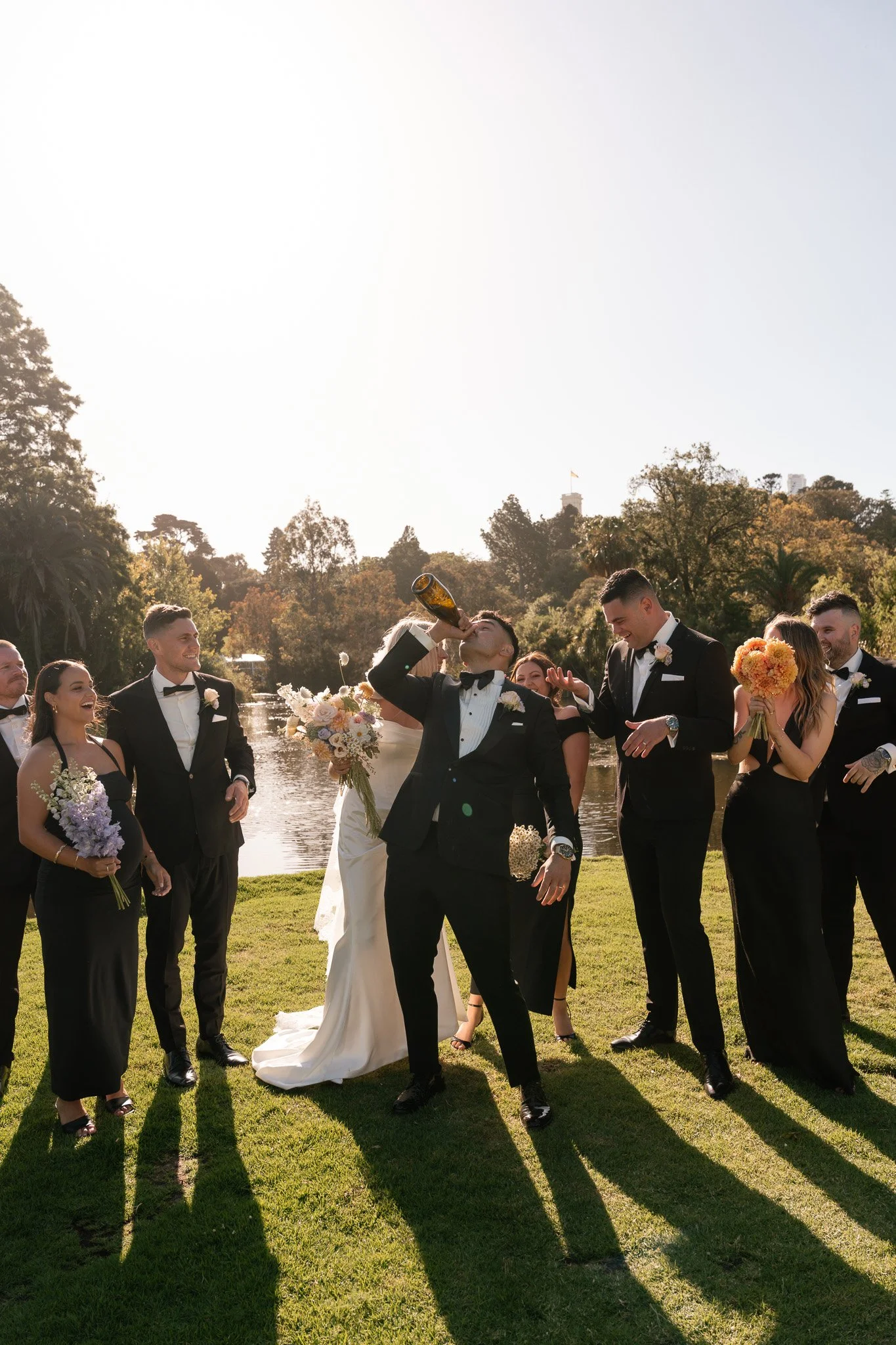 A group of wedding guests dressed in black tuxedos and gowns, celebrating by a river with a groom drinking from a bottle while others cheer and hold bouquets, during daytime.