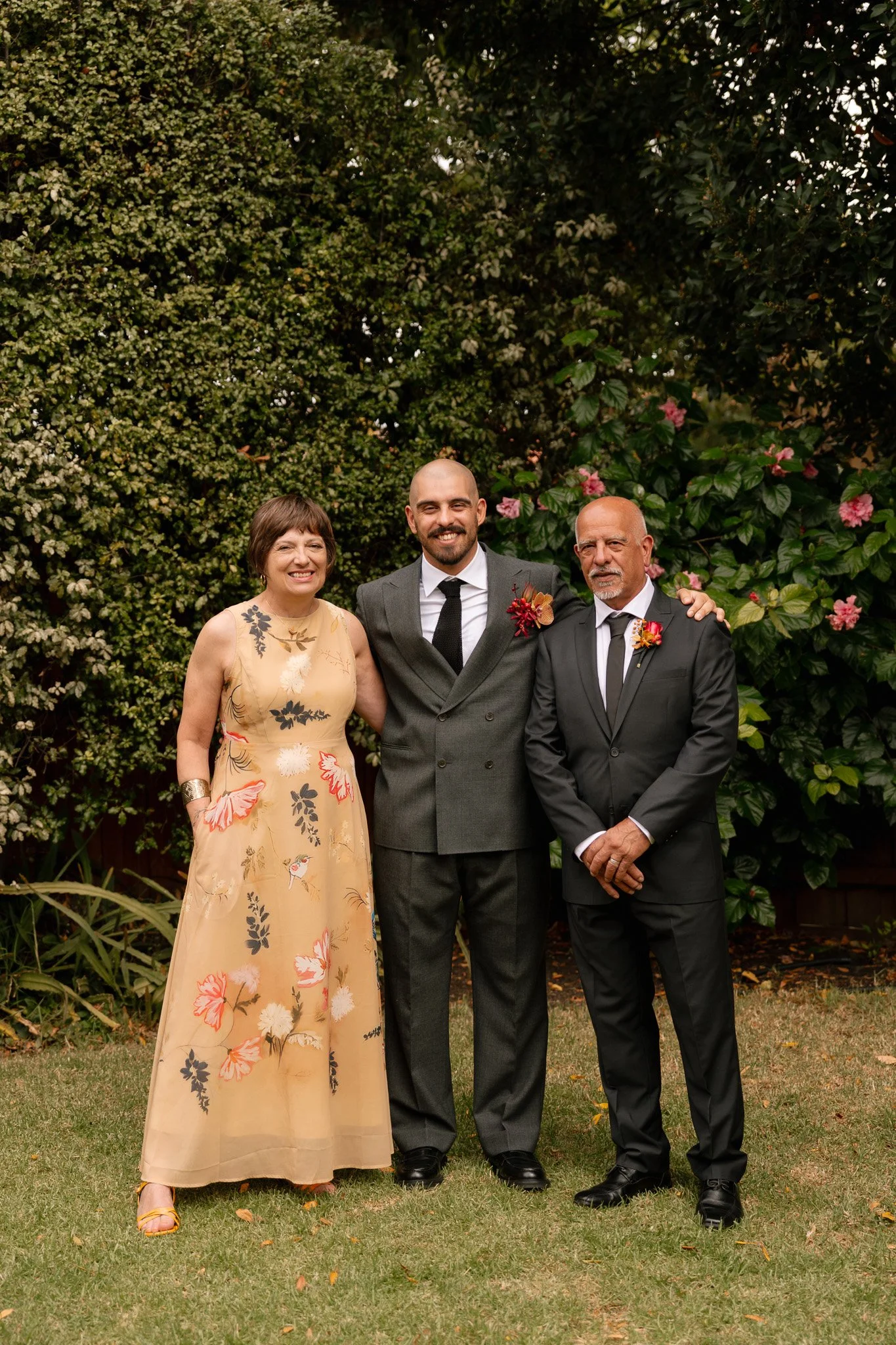 A woman in a long floral dress, a man in a dark suit, and another man in a suit with a boutonniere, standing outdoors on grass with shrubs and flowers behind them.