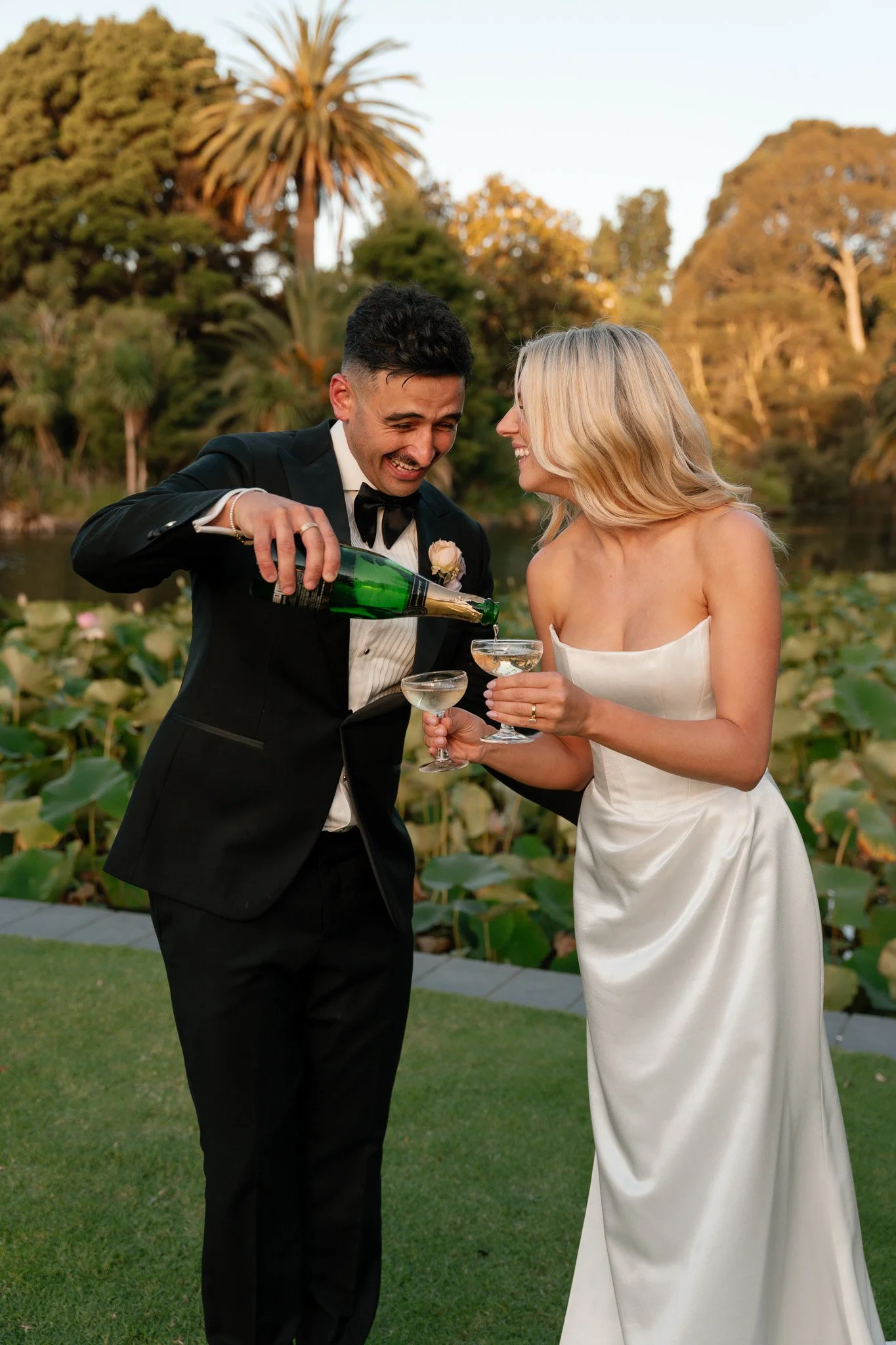 A newlywed couple celebrating with champagne outdoors near a pond with green lily pads and trees in the background.
