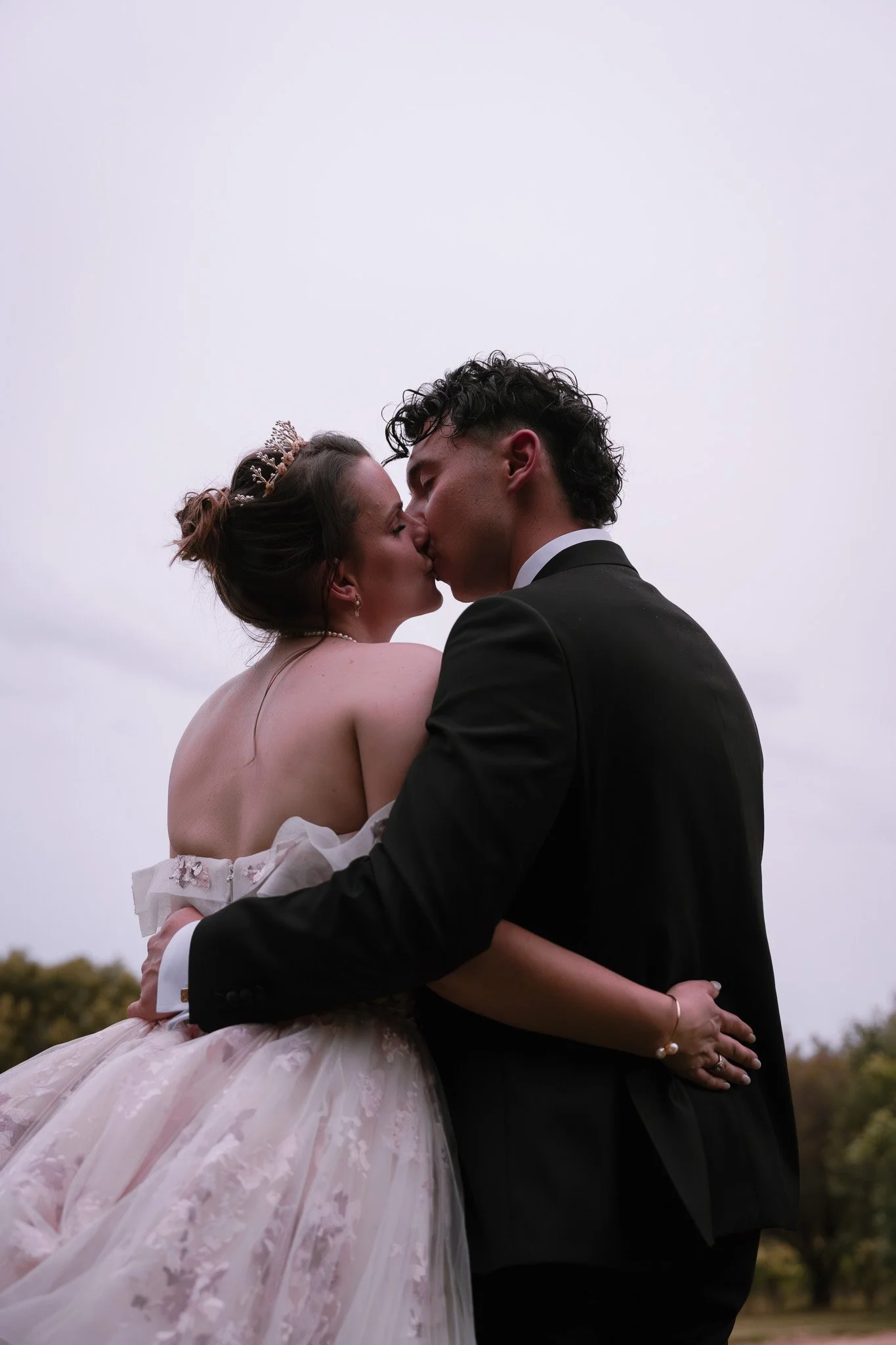 Couple in wedding attire sharing a kiss outdoors on a cloudy day, with the woman in a wedding gown and the man in a black suit.