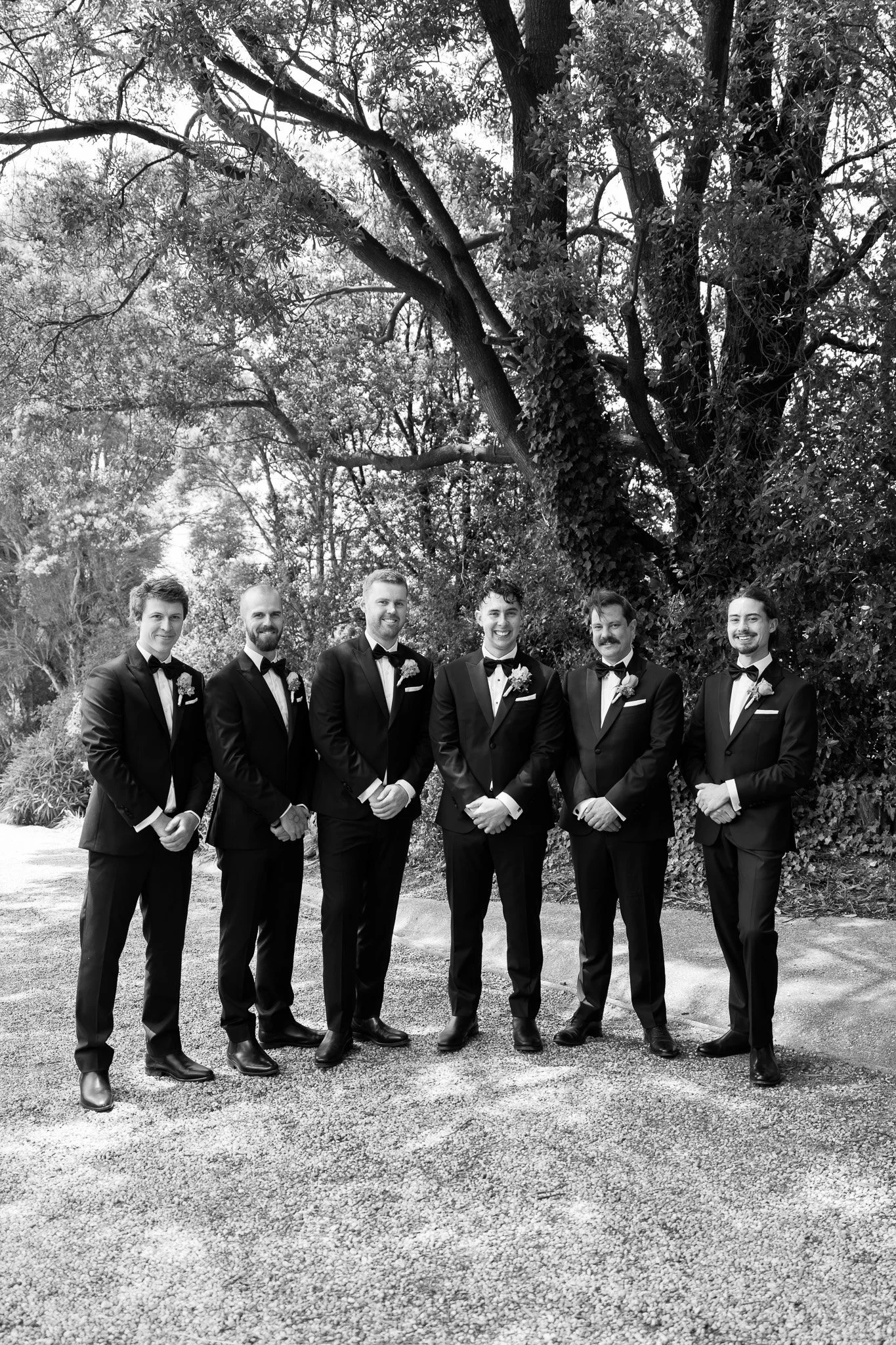 A group of six men dressed in tuxedos with bow ties, standing outdoors in a line with trees and foliage in the background.