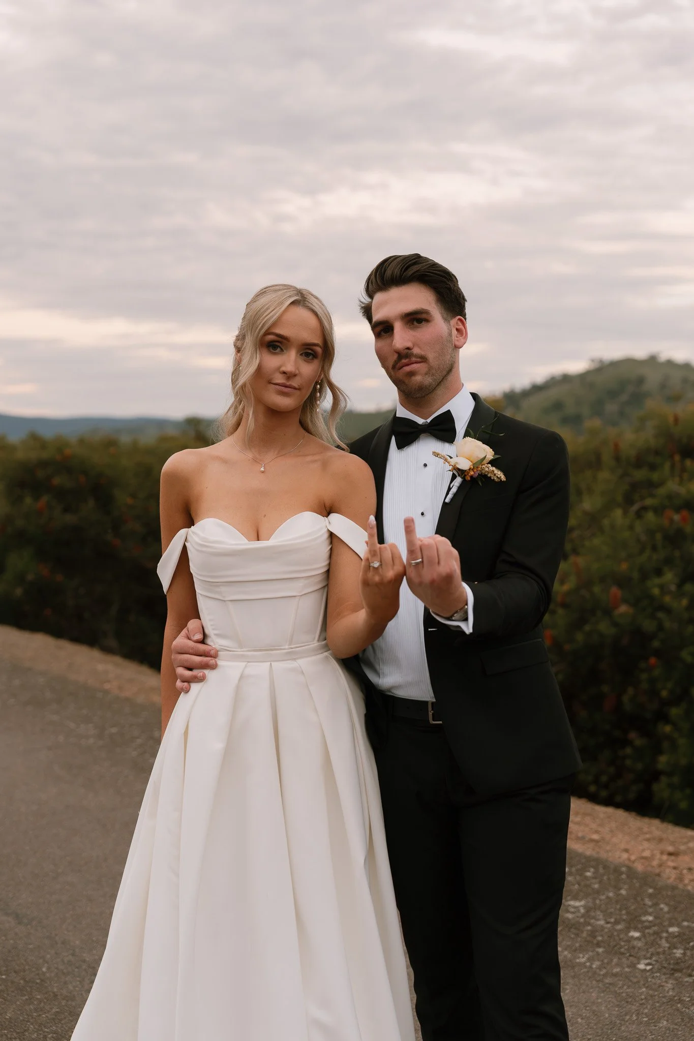A bride and groom standing outside on a cloudy day, with the bride in a white wedding dress and the groom in a black tuxedo. The groom is showing a middle finger gesture.
