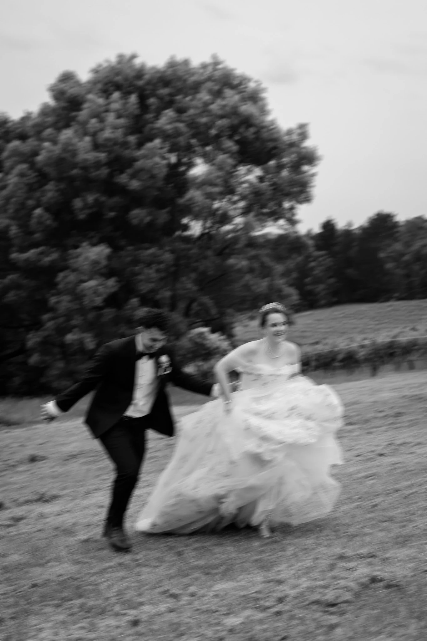 A black and white photograph of a bride and groom running outdoors, wedding dress and tuxedo, holding hands, with trees in the background.