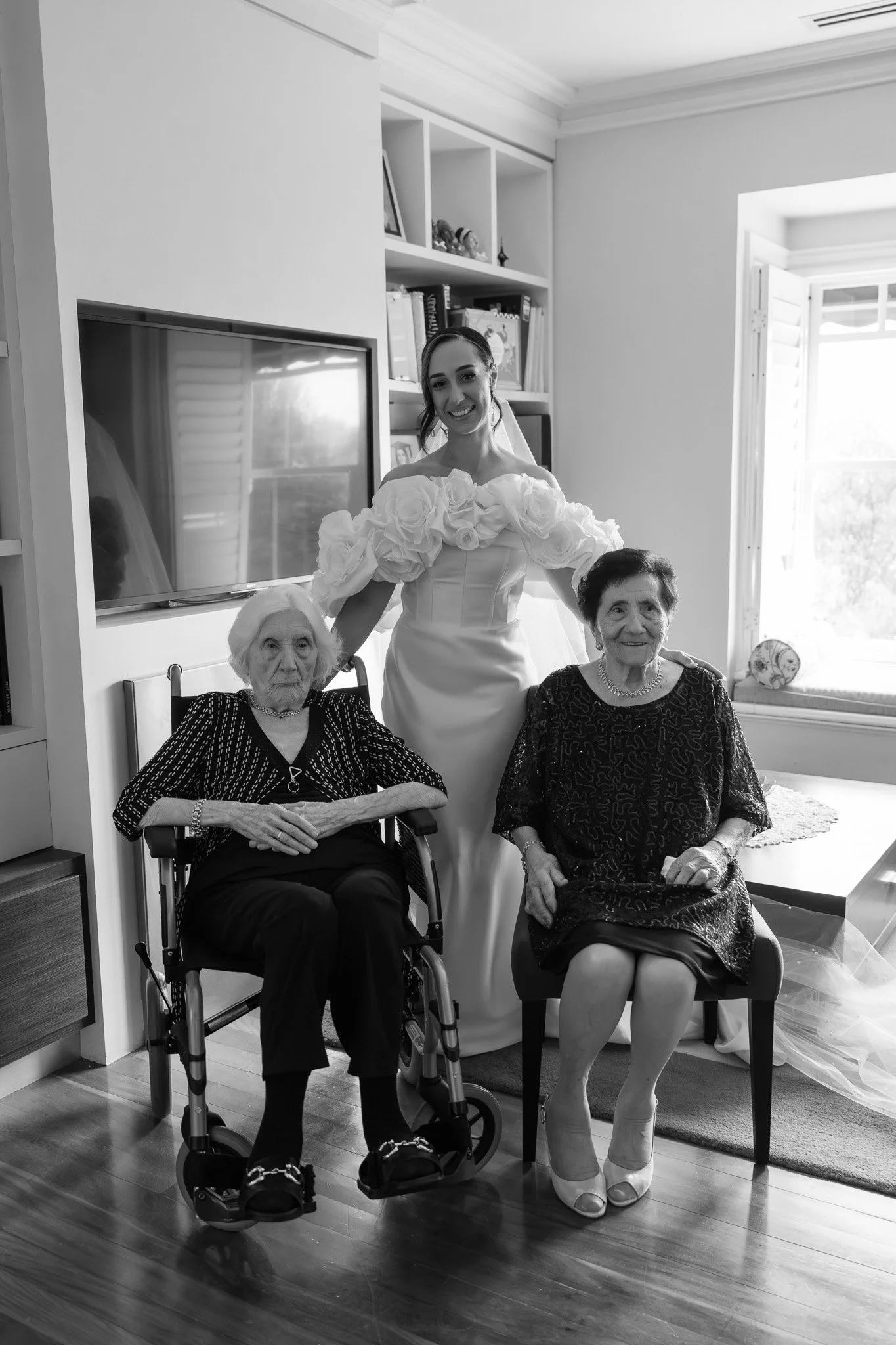 A bride in a wedding dress standing behind two seated elderly women, one in a wheelchair and the other on a chair, inside a well-lit room with a large window.