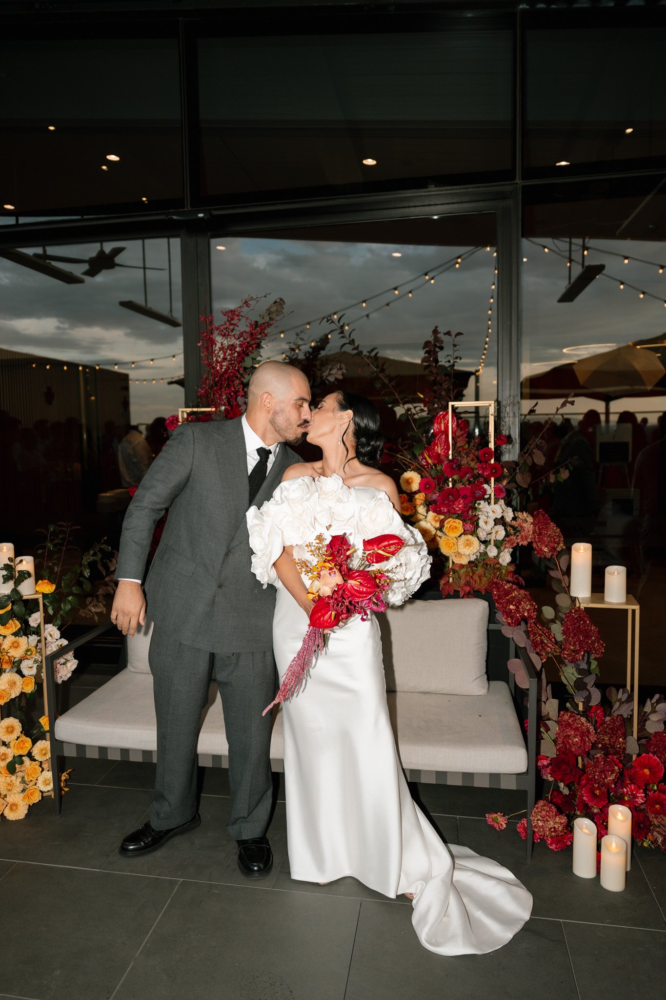 A bride and groom sharing a kiss at their wedding reception, surrounded by floral arrangements and candles.