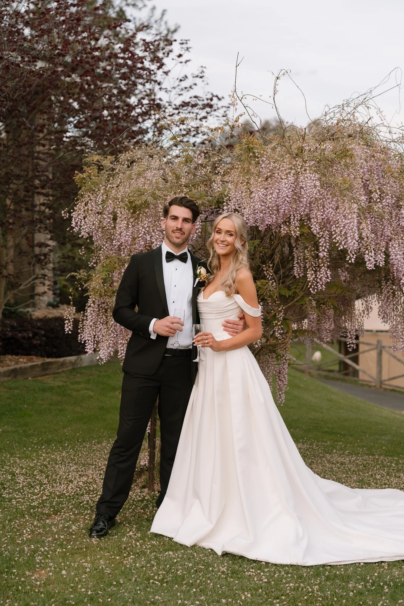 A newlywed couple in wedding attire standing outdoors in front of a flowering tree, smiling and holding champagne flutes.