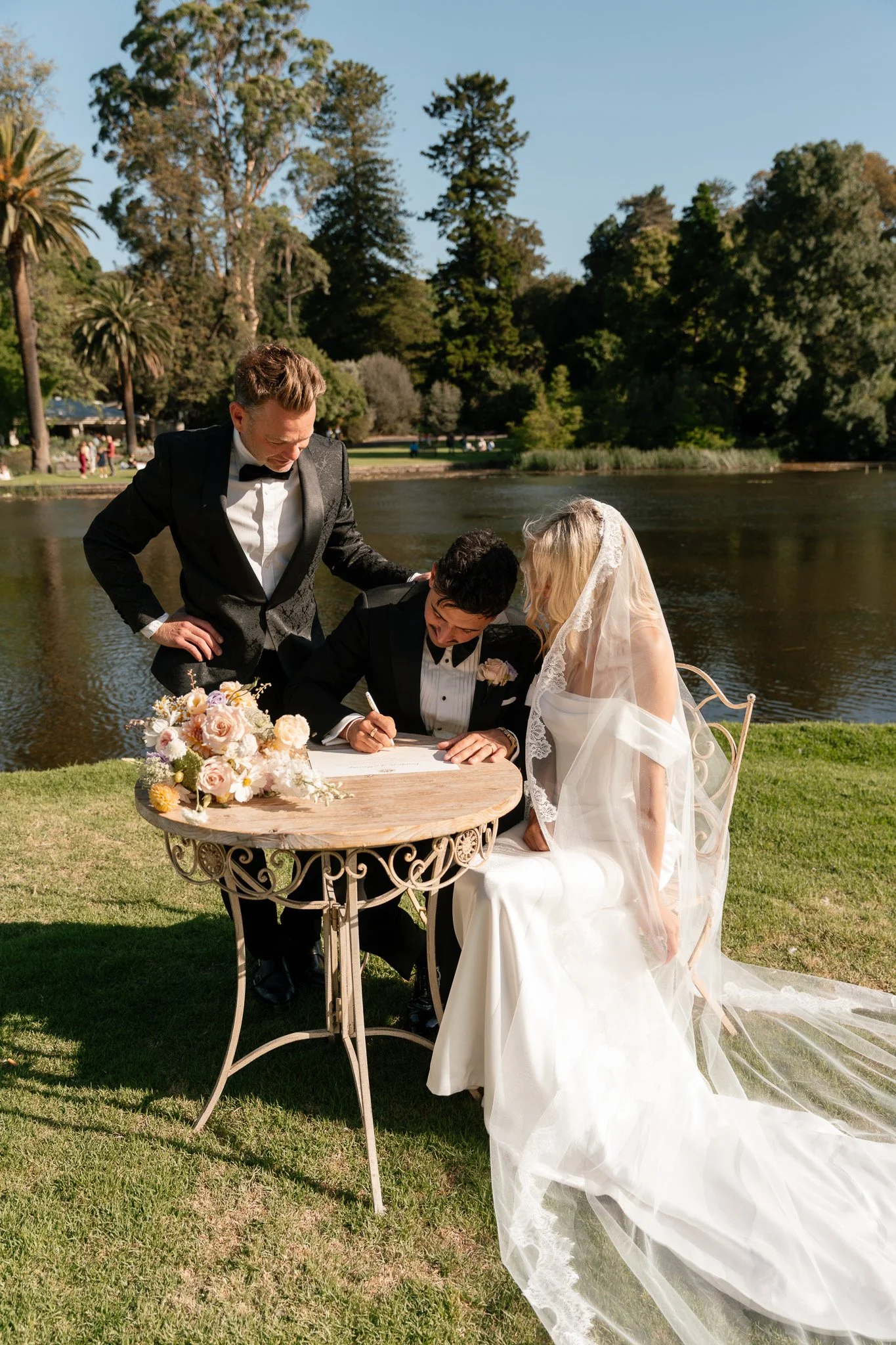 A bride and groom signing a marriage certificate outdoors next to a lake, with a man standing beside them helping them sign, and a bouquet of flowers on the table.