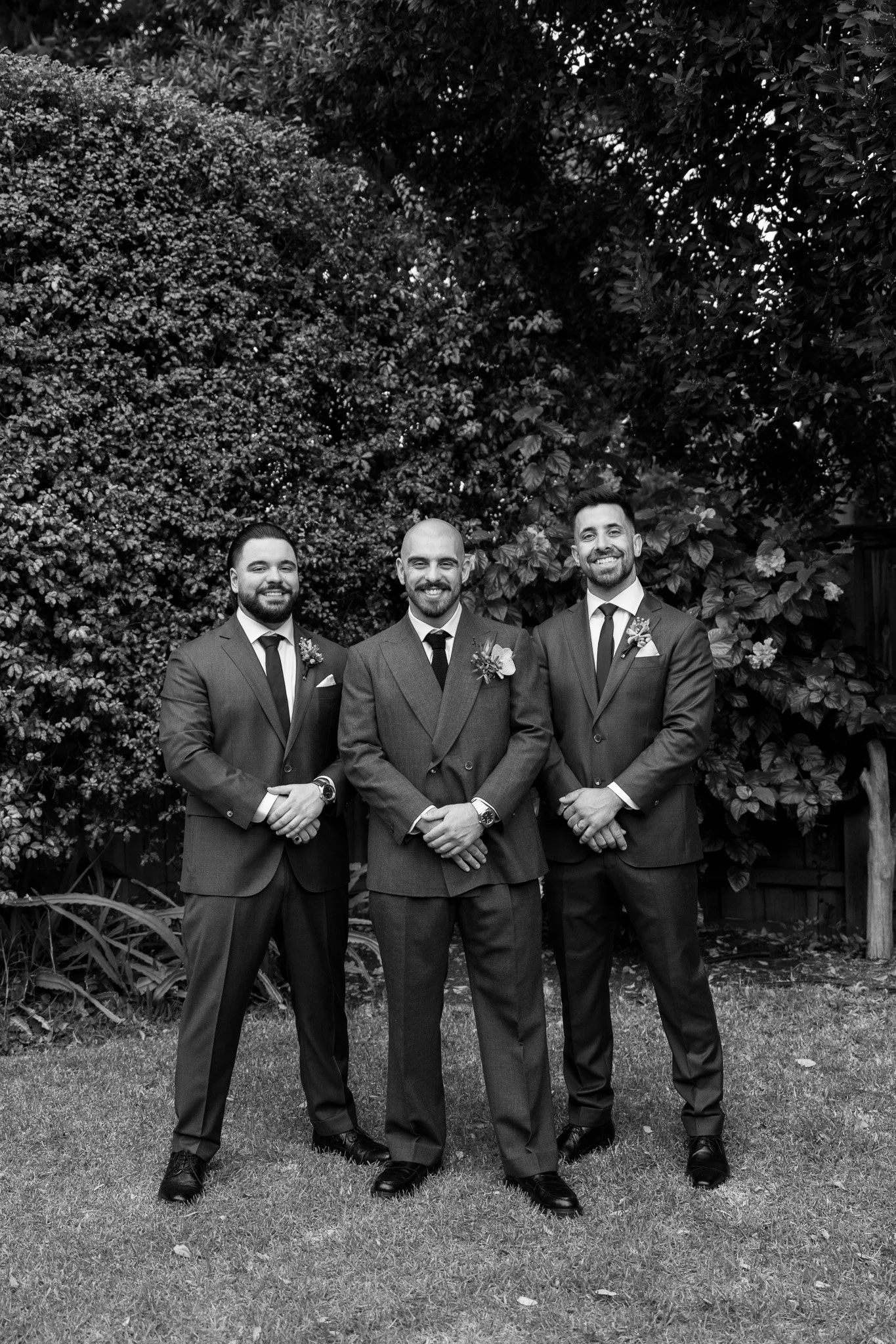 Black and white photograph of three men in suits standing outdoors in front of bushes, smiling, with the man in the middle wearing a boutonniere.