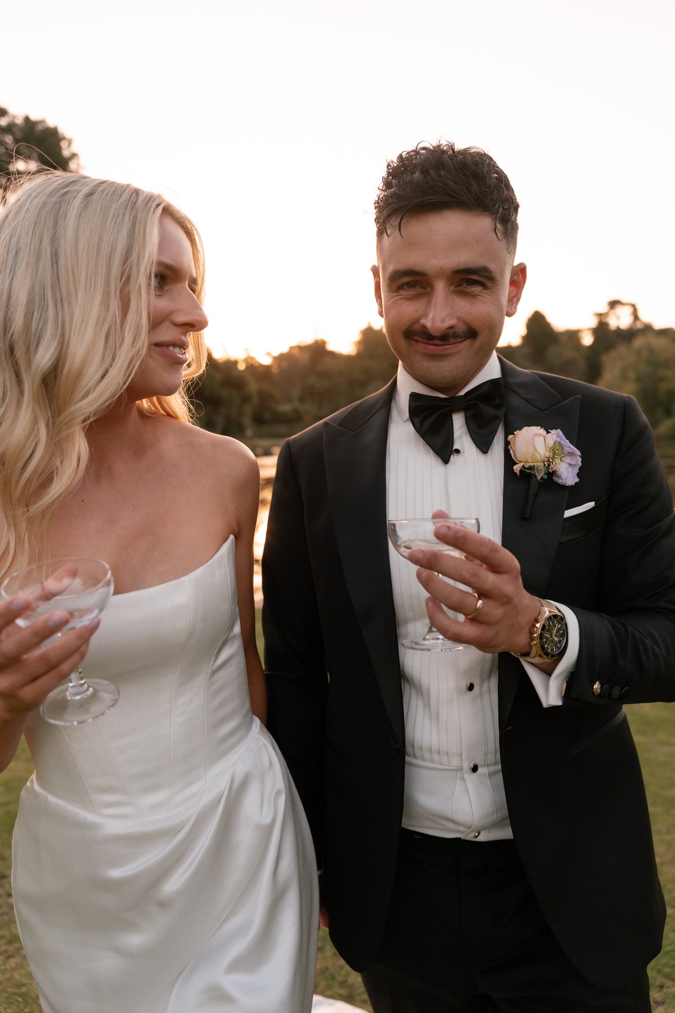 A newlywed couple at an outdoor wedding reception, holding champagne glasses, with a scenic sunset and lake in the background.
