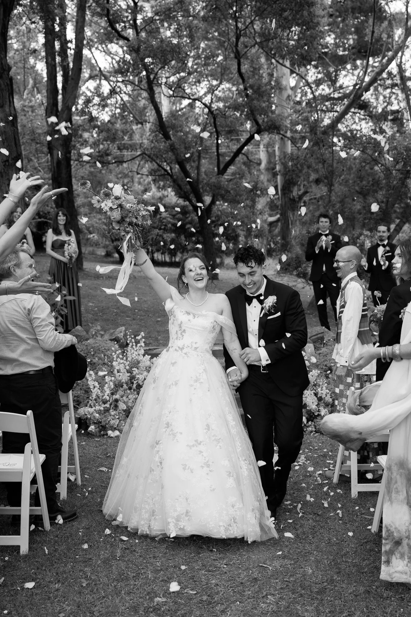 A black-and-white photo of a bride and groom walking down the aisle outdoors, smiling joyfully, as guests throw flower petals in celebration.
