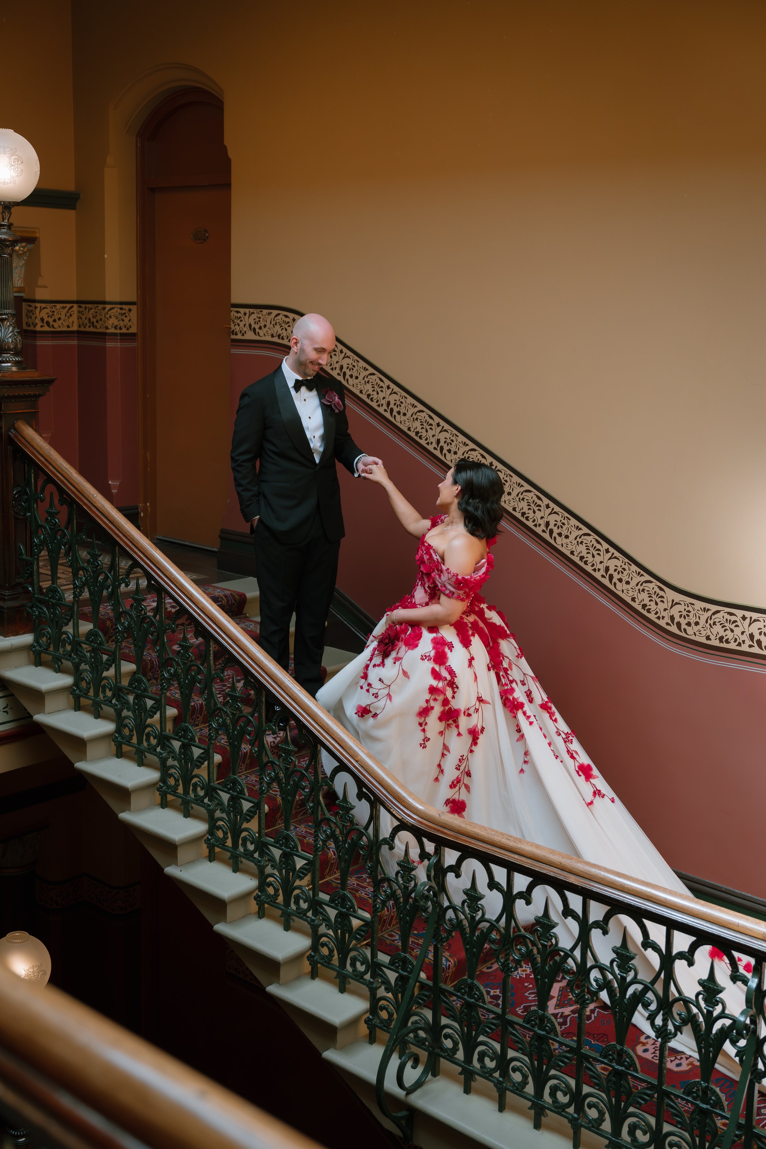 A man in a black tuxedo and bow tie holding hands with a woman in an elaborate red and white gown on a staircase with ornate wrought iron railing and decorative wall trim.