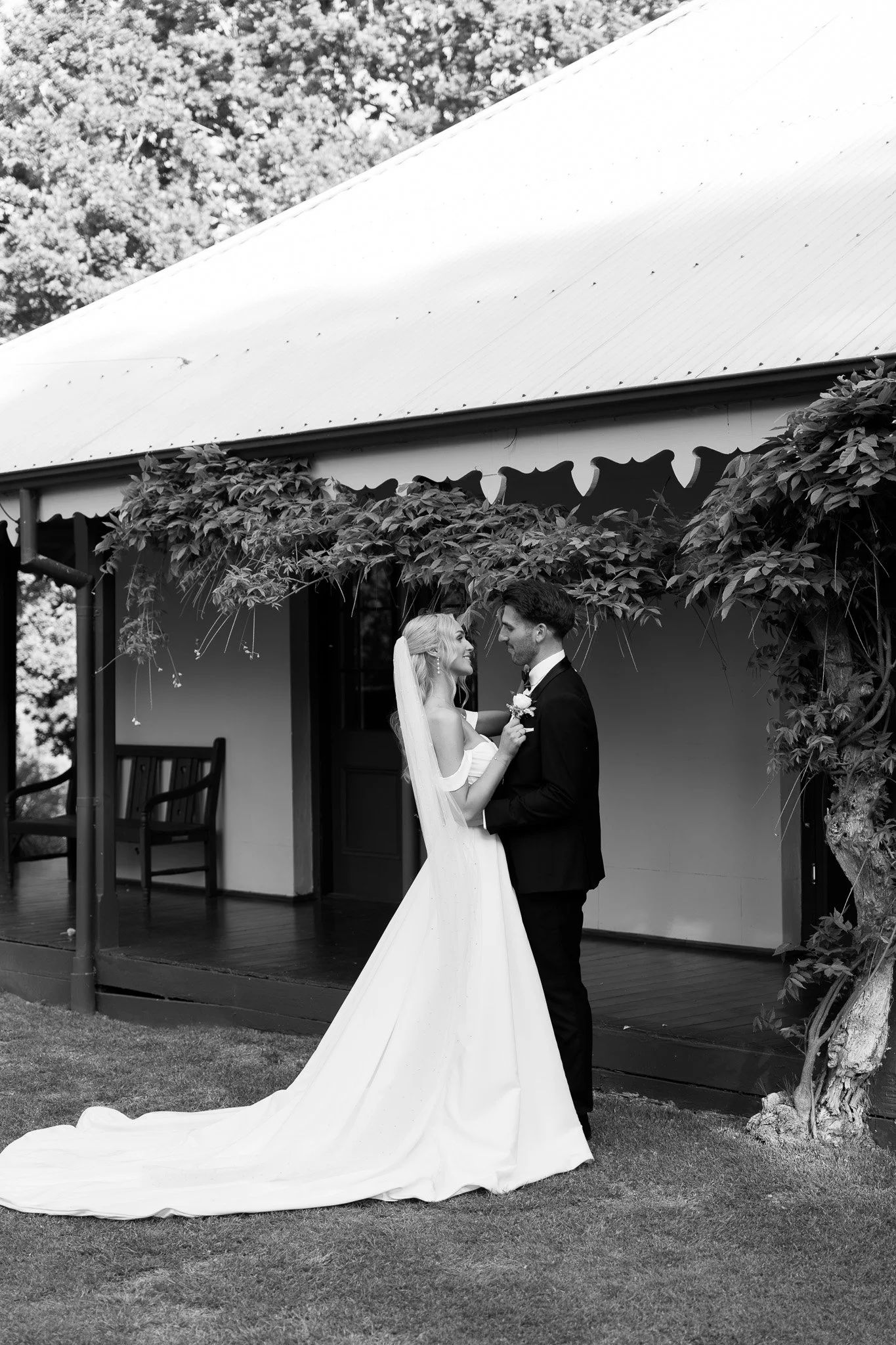 A bride and groom stand close together outside a building, smiling at each other on their wedding day. The bride is wearing a strapless white wedding gown with a long train and veil, and the groom is in a black tuxedo. The scene includes a porch with