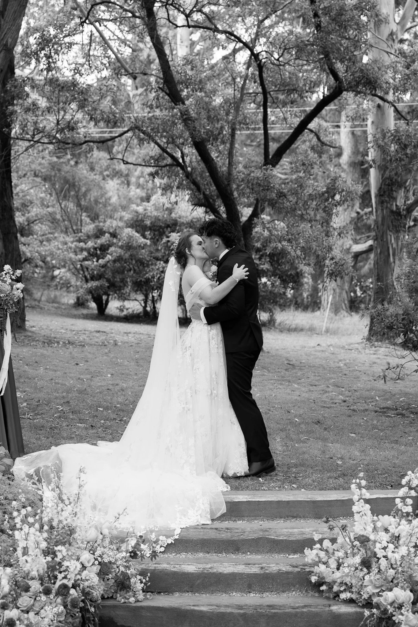 Black and white photo of a bride and groom kissing during their outdoor wedding ceremony, surrounded by trees and floral arrangements.