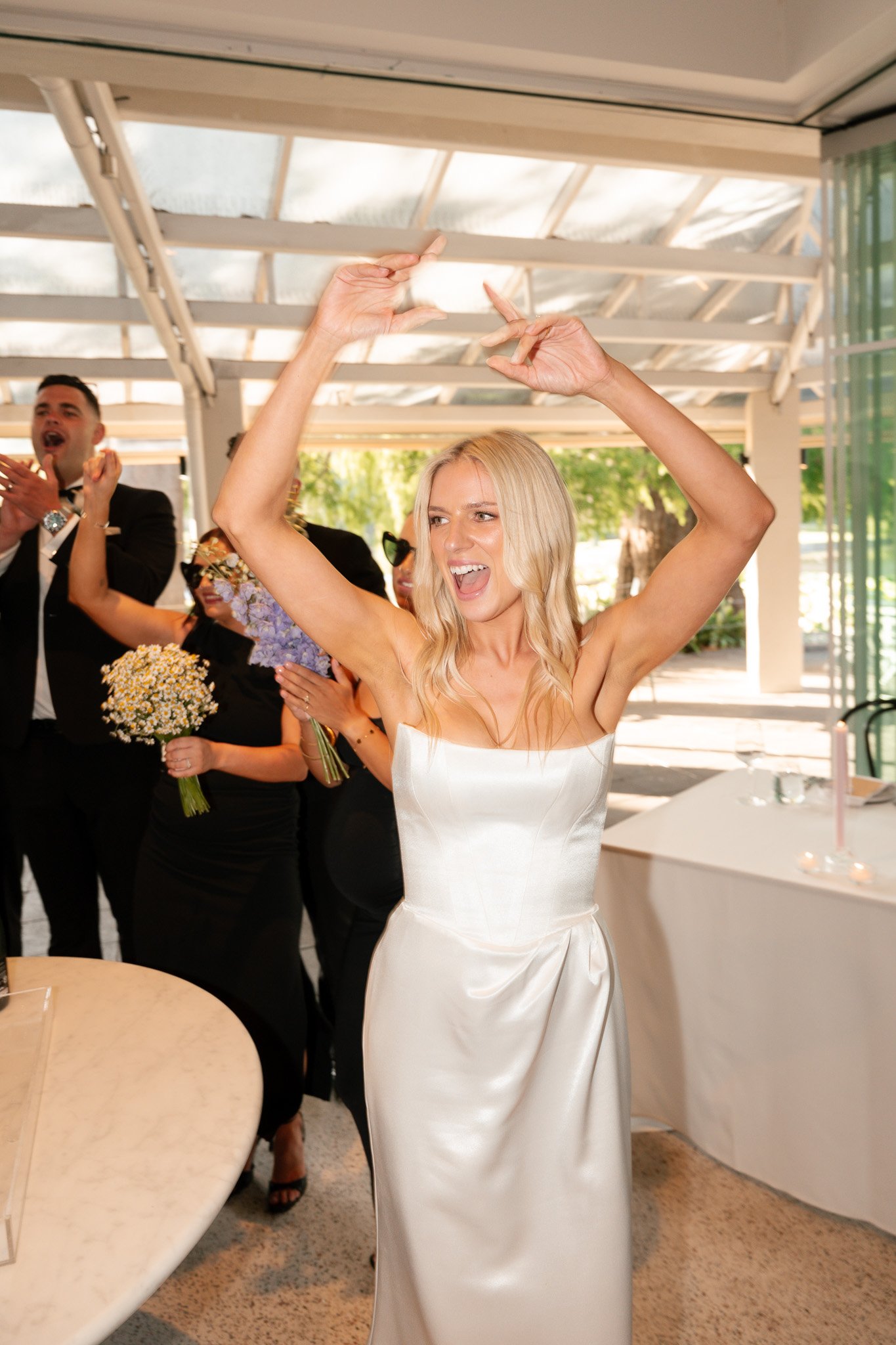 A woman in a white wedding dress dancing with her arms raised at a wedding reception, surrounded by guests in black attire, some holding bouquets, under a glass ceiling.