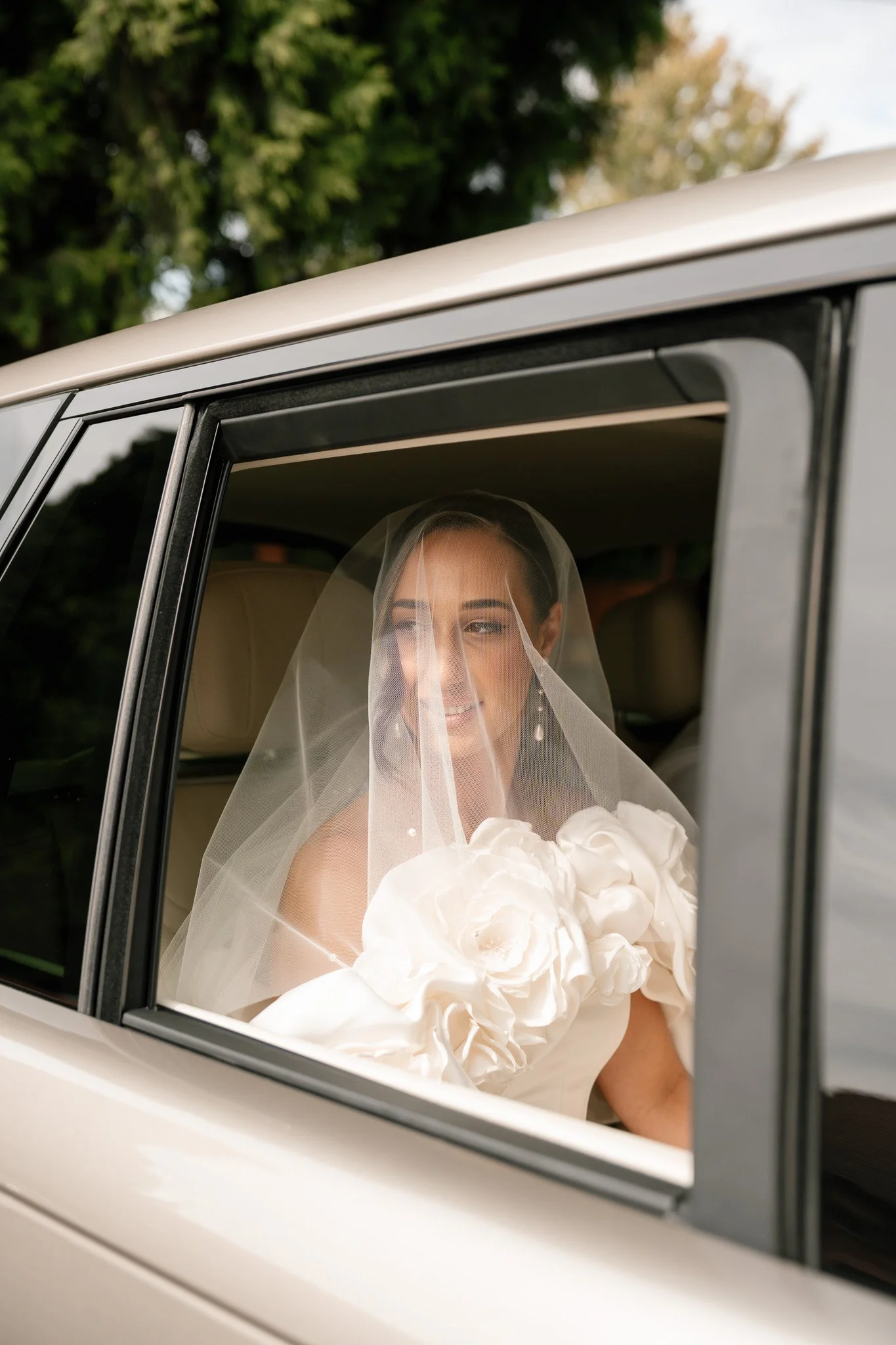 Bride sitting in car, smiling, wearing a wedding dress, veil, and earrings, looking out the window, with trees in the background.