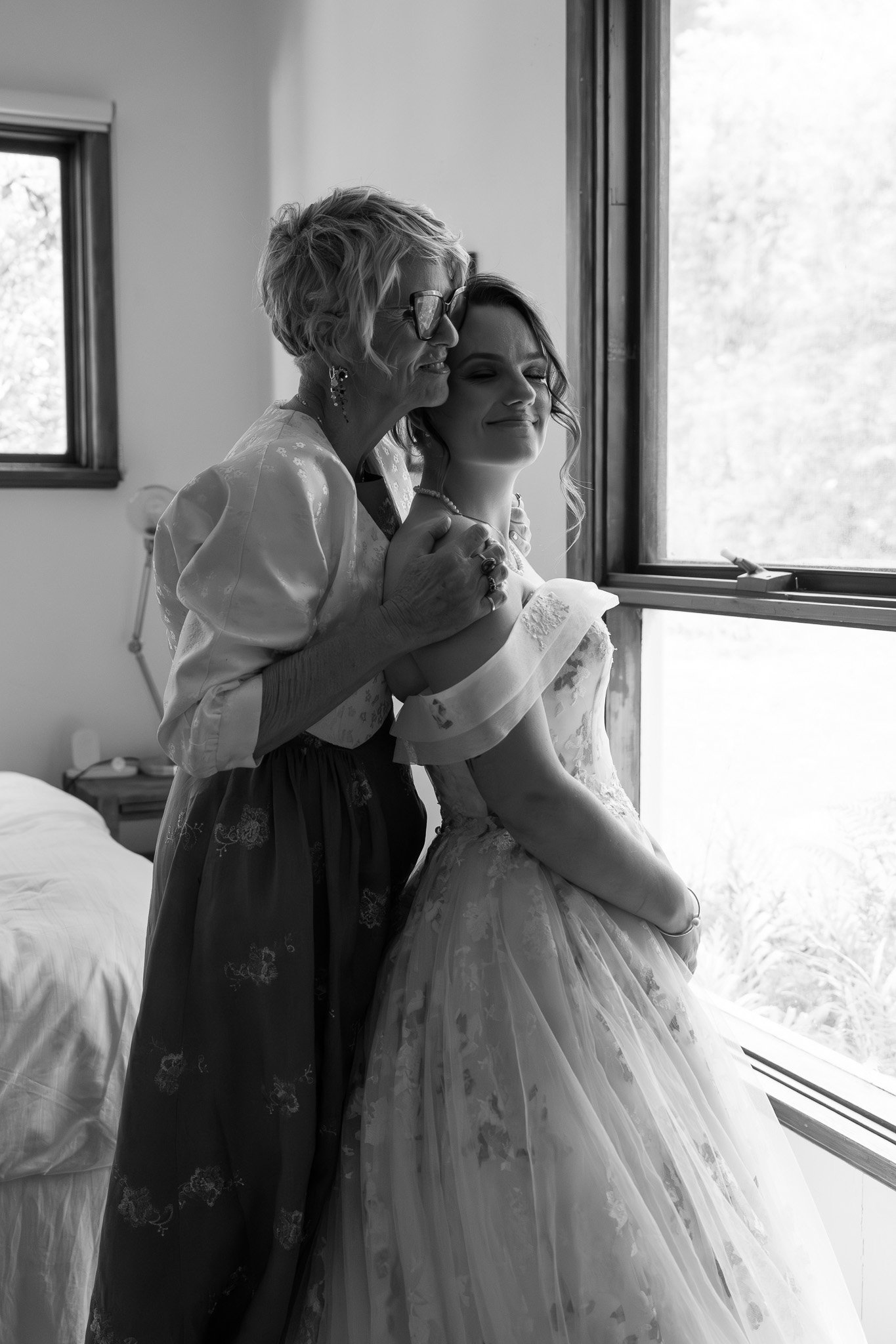 A woman in a wedding dress and an older woman, possibly her mother or grandmother, sharing an emotional moment by a window, with sunlight coming in, in a bedroom.