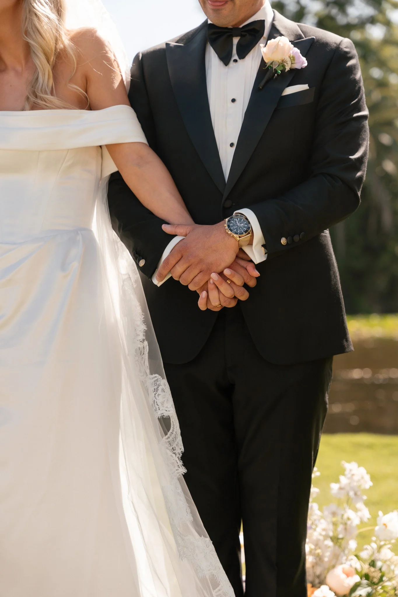 A bride and groom holding hands during their wedding ceremony. The bride is wearing a strapless white wedding gown with lace details, and the groom is in a black tuxedo with a white shirt, black bow tie, and a boutonnière. They are outdoors with flow