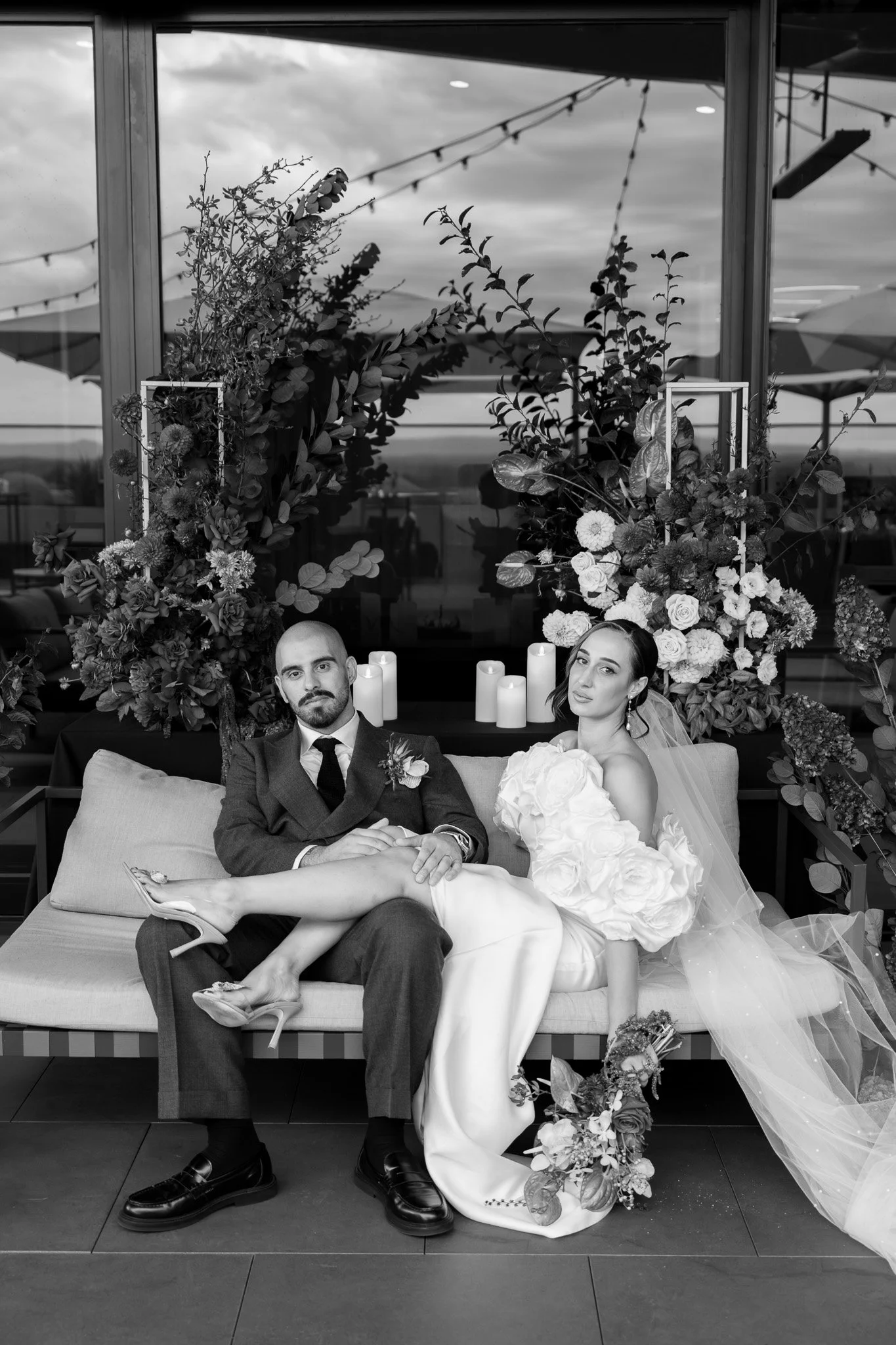 Black and white photograph of a newly married couple seated on a bench, surrounded by floral arrangements and candles, with a large window and outdoor umbrellas in the background.