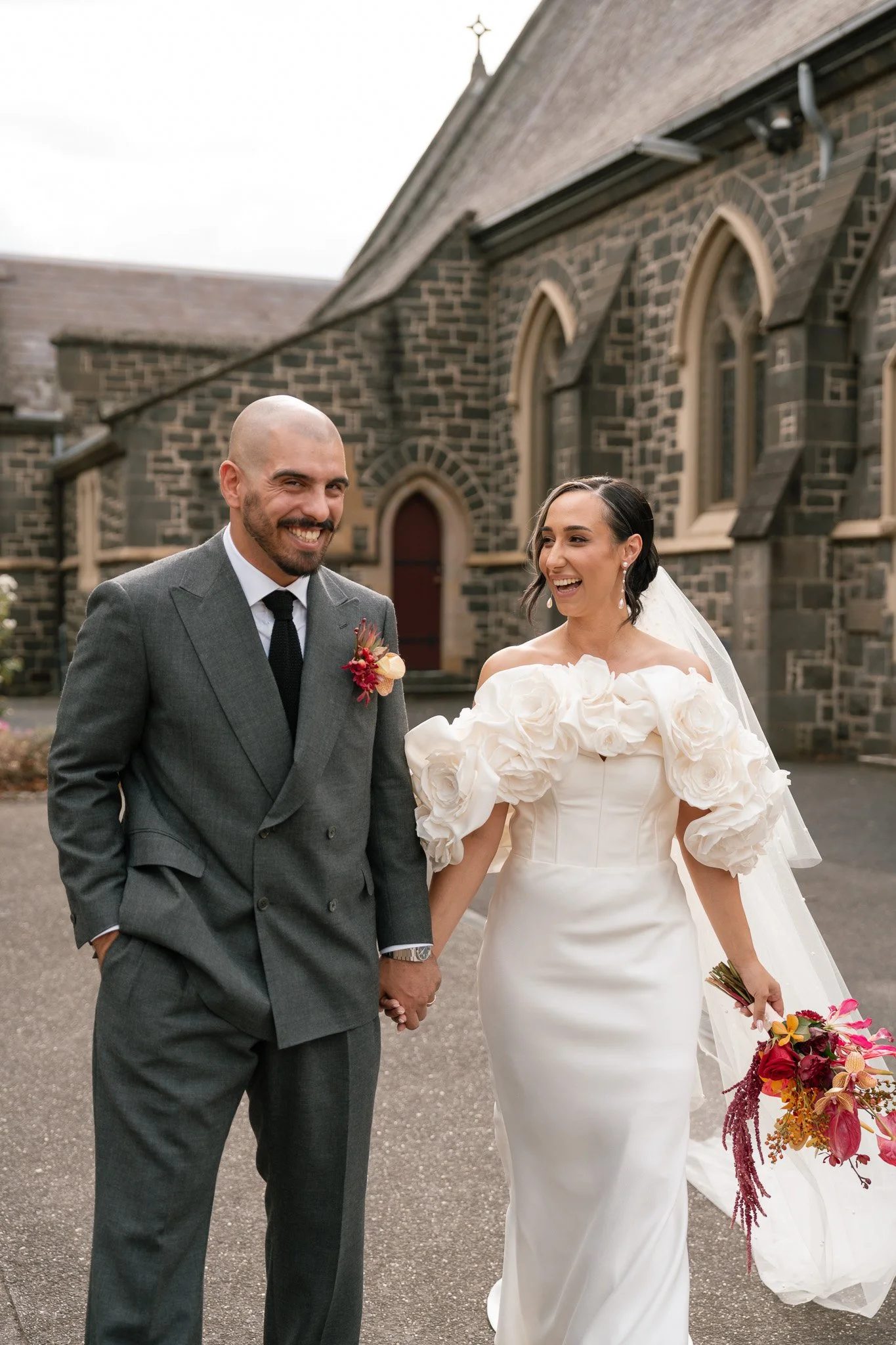 A bride and groom holding hands outside a stone church, smiling and looking happy on their wedding day.