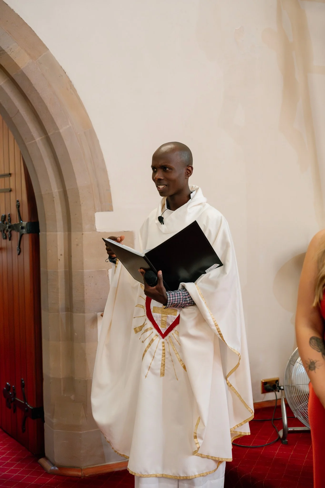 A man in a white religious robe holding a black folder and a microphone, standing inside a church or chapel with a stone archway and wooden door behind him.